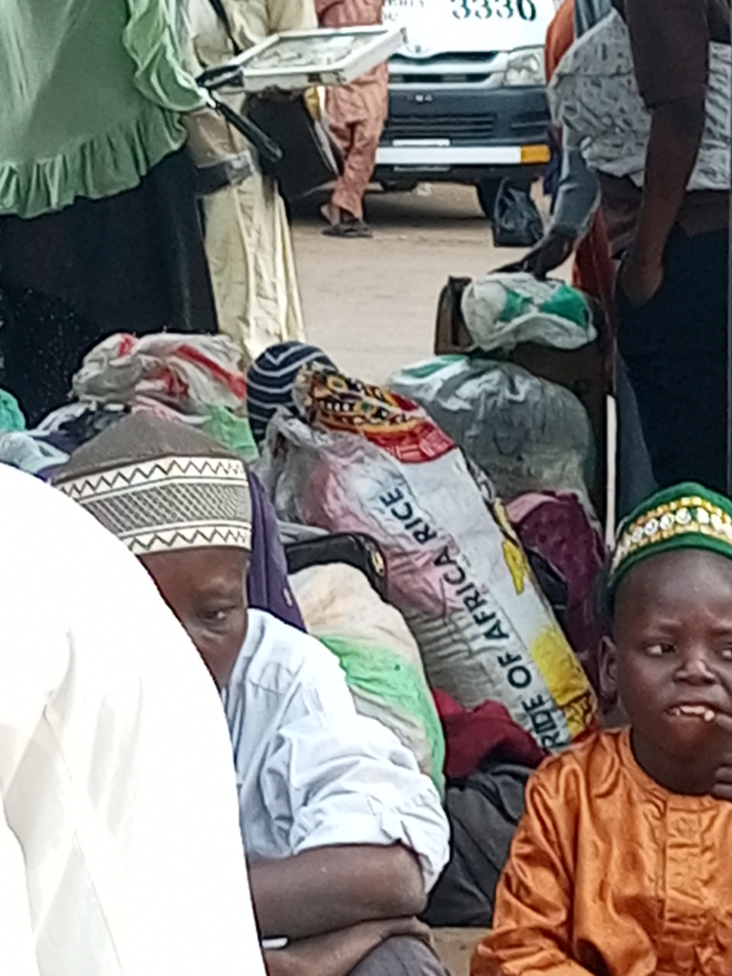 In Gombe, Nigeria, a group of individuals are gathered near a white utility vehicle displaying the number "3330". The scene appears to be a street or market area. In the foreground, two young boys are visible. One boy, on the left, is partially obscured by a white garment and is wearing a light blue shirt and a patterned kufi. The other boy, on the right, is wearing an orange kaftan and a green kufi adorned with rhinestones. He has his hand near his mouth. Behind the boys, several bags and bundles of goods are piled up. One prominent bag is labeled "RIDE OF AFRICA RICE" in large, bold letters. Other individuals in the background are dressed in traditional clothing, including a woman in a green headscarf and dress, and another person in a cream-colored robe. The overall impression is of a busy, outdoor environment, possibly related to commerce or travel.