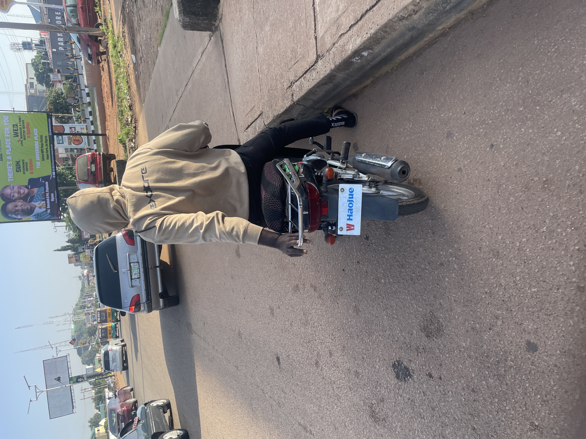 The image captures a scene on a road in Jos, Nigeria, likely during the day. In the foreground, a person wearing a light brown hooded sweatshirt and black pants is seated on a Haojue motorcycle. The motorcycle has a license plate with blue and white lettering that appears to read "W HAOJUE". The rider's left hand is visible resting on the motorcycle's grab bar. The background shows a busy street with various vehicles, including cars, a tuk-tuk, and other motorcycles. On the right side of the road, a large billboard advertising a church service is visible, with details about Sunday and Wednesday services and the location as "The Governing Place, Behind NNPC Mega Station, Dogon Karta, Jos Nigeria". Further in the background, buildings and streetlights are present, indicating an urban environment. The road surface is asphalt, with some patches of dirt and debris along the side. The overall impression is of a typical street scene in a Nigerian city.