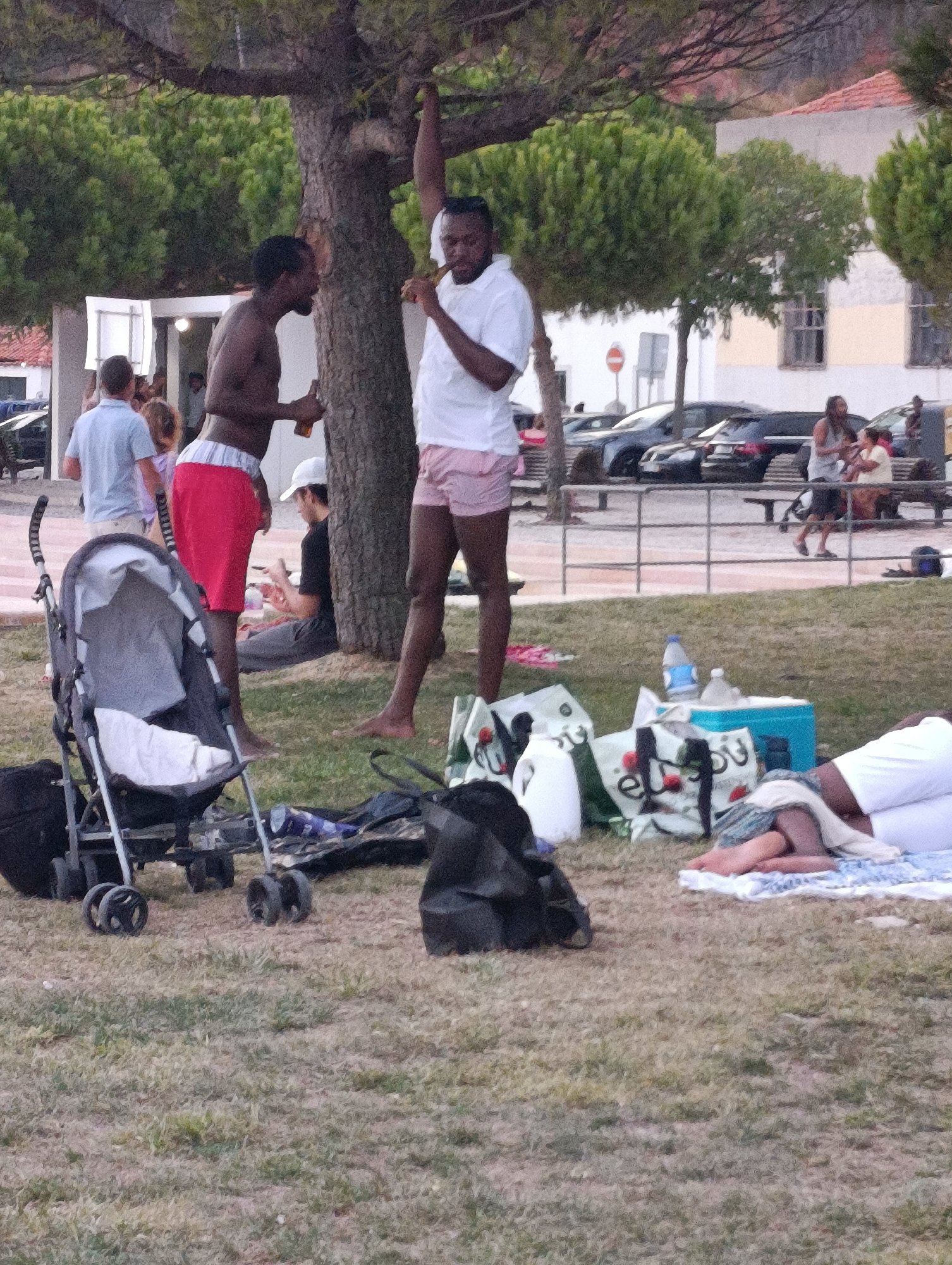 This image depicts an outdoor scene, possibly a park or recreational area, with people gathered. In the foreground, a stroller is visible on grassy ground. Further back, several individuals are standing near a tree. One person, wearing a white shirt and pink shorts, is holding a bottle. Another person, bare-chested and wearing red shorts, is also near the tree. In the background, there are parked cars and buildings, suggesting an urban or semi-urban environment. The overall atmosphere appears casual, with people socializing and relaxing outdoors. The location is identified as being in the city of Setúbal, Portugal, near the waterfront, as evidenced by the presence of cars typical of urban parking areas and buildings that align with Portuguese architectural styles.