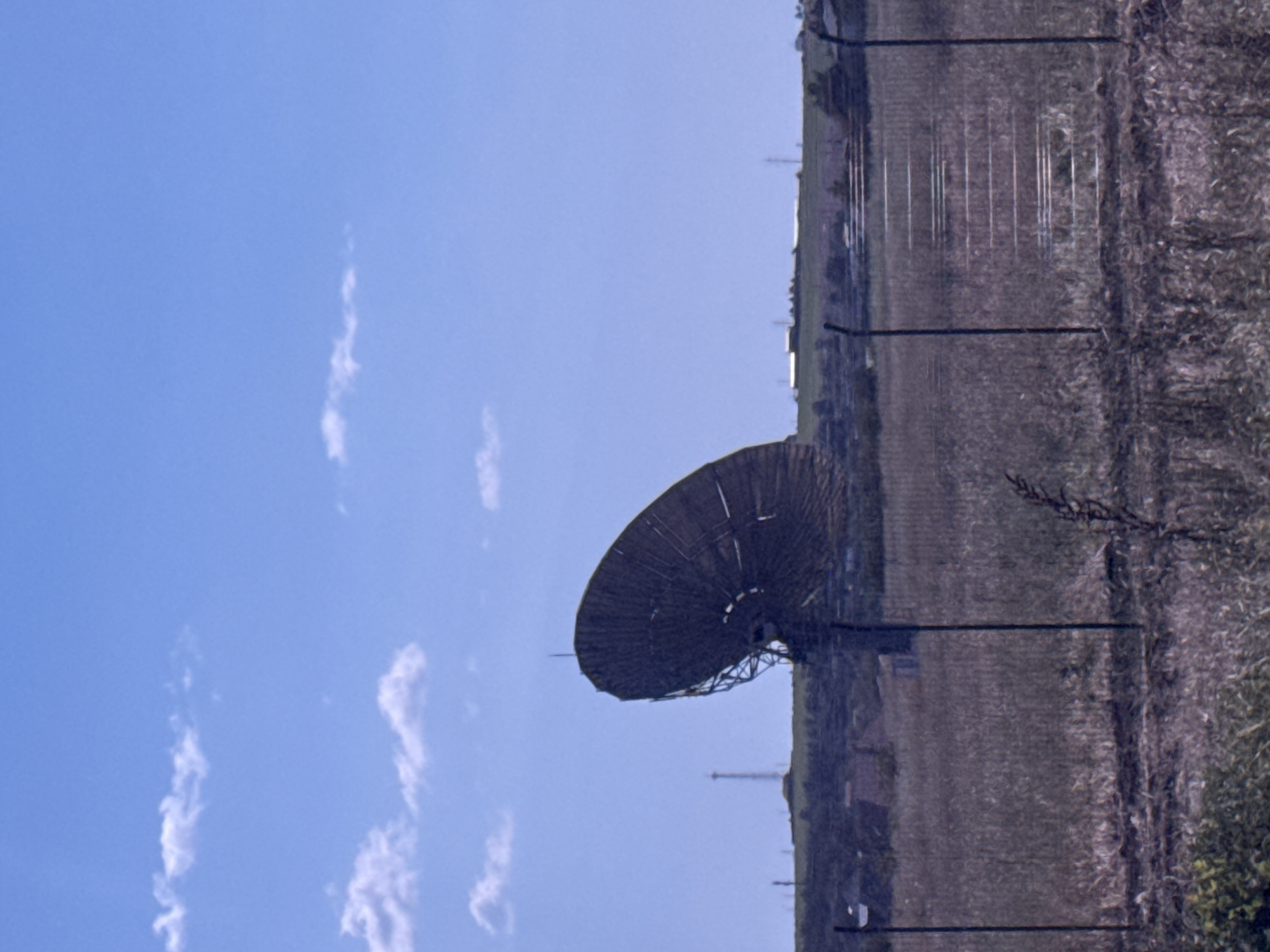 A large, dark satellite dish stands in a field under a bright blue sky with scattered white clouds. The dish is positioned to the right of the frame and is angled upwards. In the background, a faint transmission tower is visible on a distant hill. The foreground consists of dry, brown grass, with a chain-link fence running horizontally across the image. The overall scene appears to be an open, rural area, possibly a communication facility.