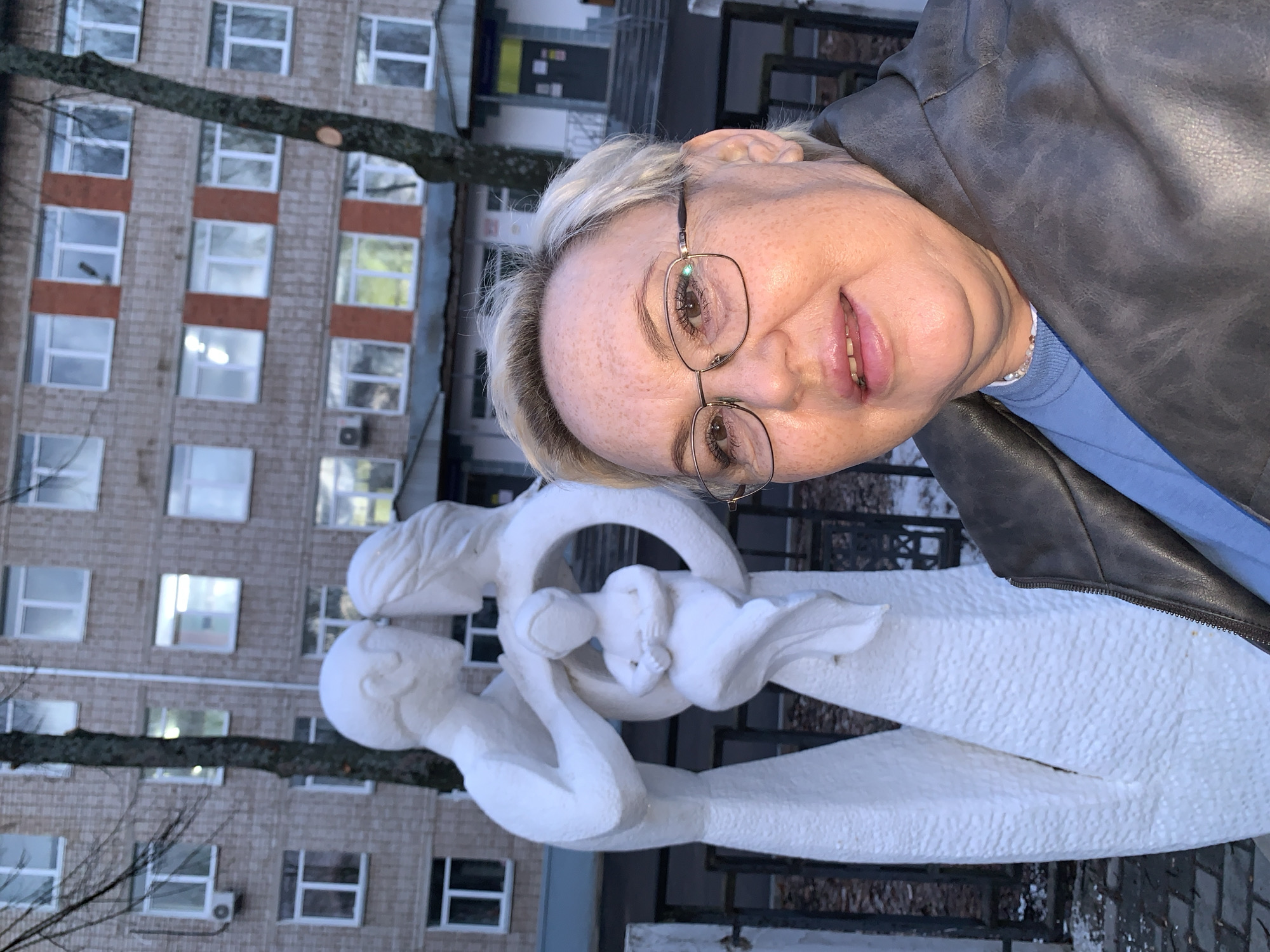 A woman with blonde hair and glasses is posing for a selfie next to a white sculpture in the city of Chaikovsky, Russia. The sculpture features stylized human figures, with one figure cradling another within a circular form. The sculpture is situated in front of a multi-story brick building with numerous windows and a tree obscuring parts of the building and the sculpture. The woman is wearing a blue shirt under a dark-colored jacket. There is a black metal fence and some vegetation near the base of the sculpture. The pavement is damp.