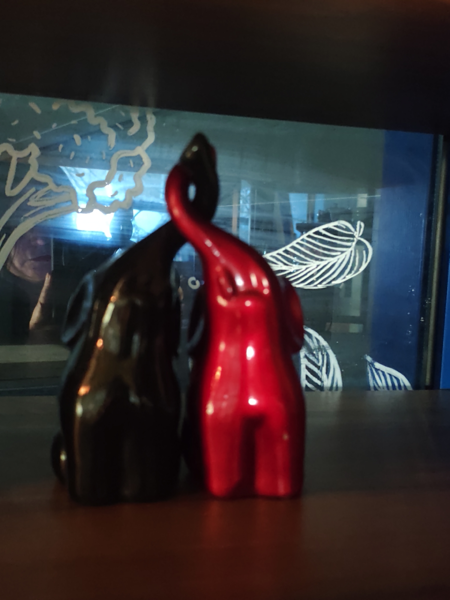 This is a close-up, slightly blurry, eye-level shot of two stylized elephant figurines, one black and one metallic red, positioned on a dark wooden surface. The figurines appear to be facing each other or a common point of interest, with their trunks curved upwards and intertwined, suggesting affection or togetherness. The background is a dark, reflective surface with white line drawings of leaves and abstract shapes, possibly a mural or a decorated window. A faint reflection of a person is visible in the background, appearing on the left side of the image. The lighting is dim and atmospheric, casting subtle highlights on the figurines. The setting does not provide any direct indicators of its location within Sochi, Russia.