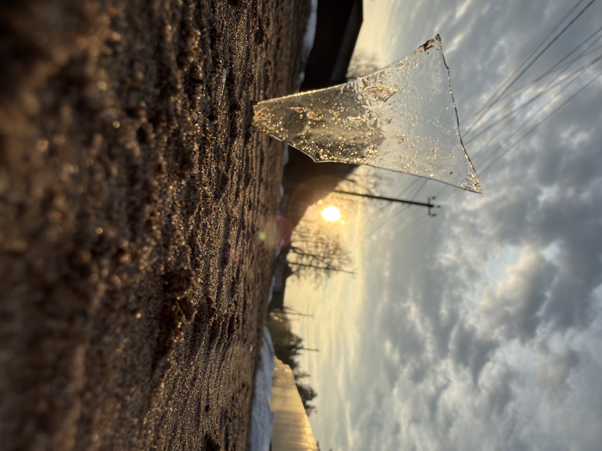 A shard of ice is perched on a dark, textured surface, likely frozen earth or soil. The ice is triangular with jagged edges and internal imperfections visible within its transparent structure. The sun is positioned low on the horizon, casting a golden glow through the ice and across the textured surface. In the background, silhouettes of bare trees and utility poles are visible against a cloudy sky. Above the tree line are the power lines running from the utility poles. The setting suggests an outdoor location, possibly at the city of Chaikovsky, Russia during late winter/early spring. No people are present in the frame.