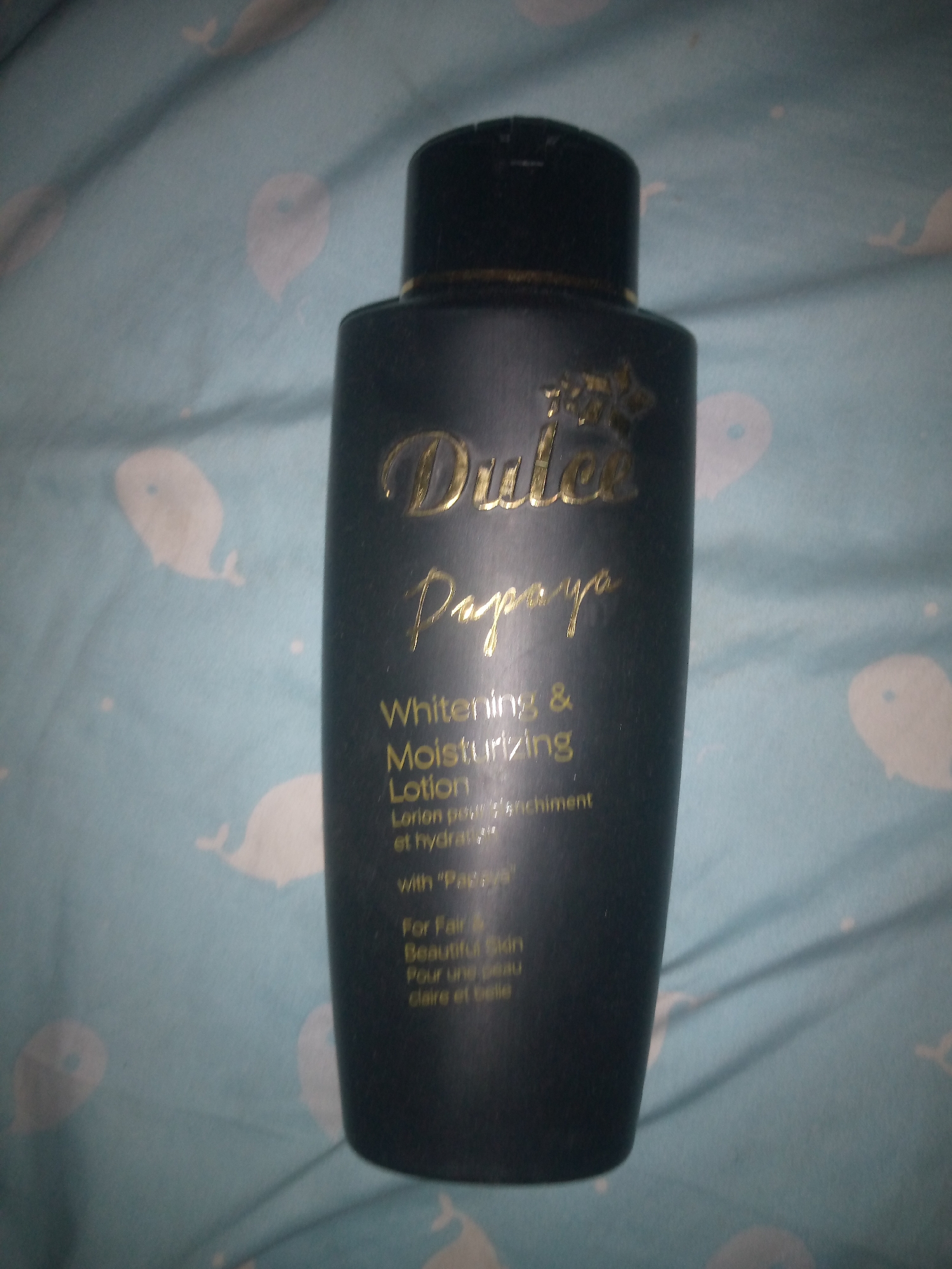The image shows a dark-colored bottle of "Dulce Papaya" whitening and moisturizing lotion. The bottle has a black cap and gold lettering. The text on the bottle indicates it is "Lotion pour peau d'enchantment et hydratante" and "For Fair & Beautiful Skin," along with French text "Pour une peau claire et belle." The bottle is placed on a light blue fabric with a pattern of white whale shapes. The image does not provide any information that would allow for the determination of its location in Jalingo, Nigeria.