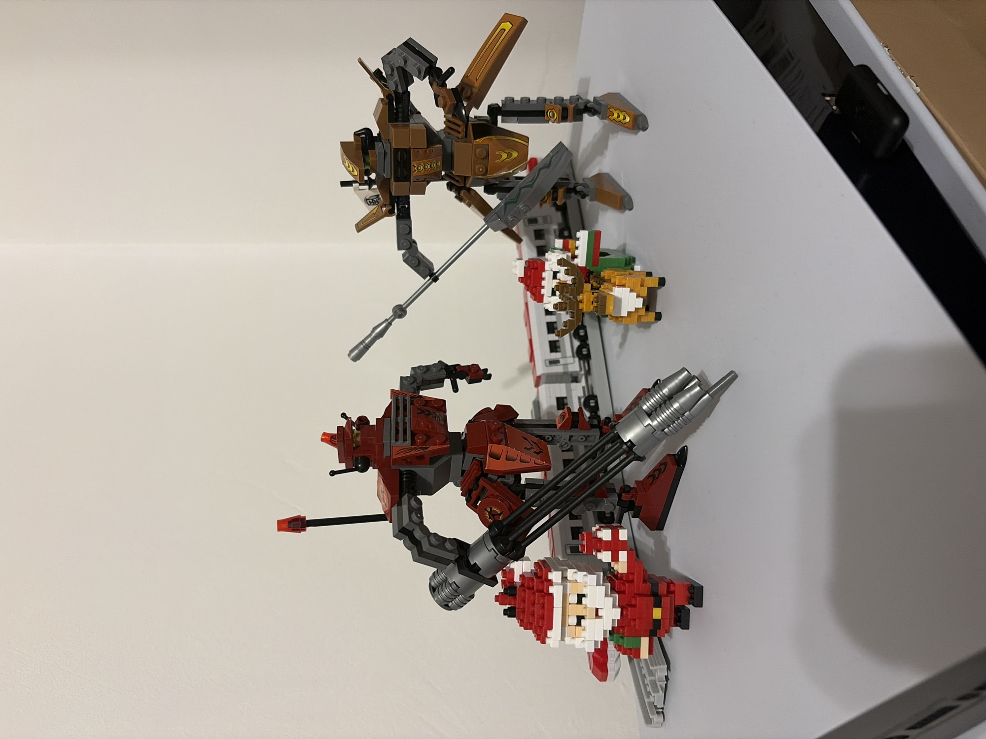 The image shows two Lego-like construction toys, arranged vertically. The top toy is primarily brown and grey, with a long, silver spear. The bottom toy is mainly red and grey, with a triple-barreled silver gun. Both toys have a pixelated Santa Claus figure attached to them. The toys are resting against a white wall and part of a white surface, likely a table or shelf. The setting is indoors. There is no indication of the location being Burgas, Bulgaria.
