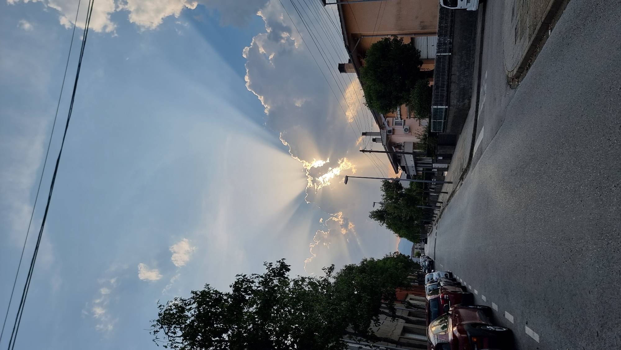 A street-level, eye-level, wide shot depicts a road lined with parked cars and trees, with houses and buildings on the right side of the street. The sky above is partially cloudy with rays of sunlight beaming through the clouds, creating a dramatic effect. The road is asphalt, with white dashed lines marking parking spaces on the left and a pedestrian crossing on the right. A few streetlights are visible along the road. The buildings on the right appear to be residential, with a variety of architectural styles and colors. The trees lining the street are lush and green, providing shade. In the distance, a mountain range can be seen. The scene is set in the town of Sant Joan Les Fonts, Spain, and appears to be during the late afternoon or early evening, given the angle of the sun and the lighting conditions. There are no people visible in the frame.