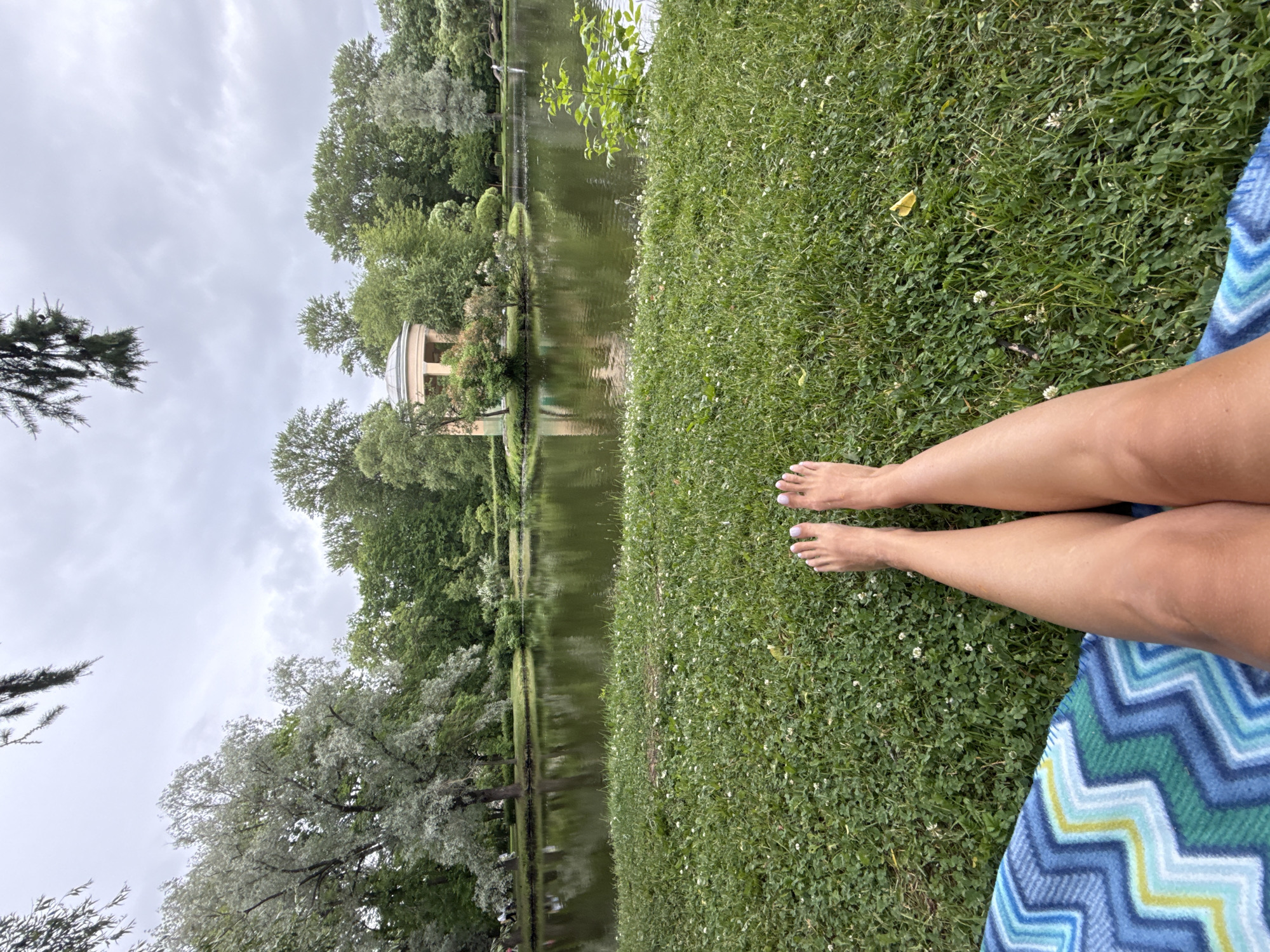 In this medium shot, a person's legs and feet are prominently displayed in the foreground, resting on a colorful striped blanket laid out on a lush green lawn. The person's bare feet, with neatly painted toenails, point upwards and slightly to the right. Beyond the lawn, a tranquil body of water reflects the overcast sky and the surrounding greenery. On the far side of the water, a picturesque scene unfolds with a variety of trees, some with silvery foliage, and a charming classical-style rotunda building with a domed roof. The overall setting appears to be a park or garden, likely on a cloudy day.