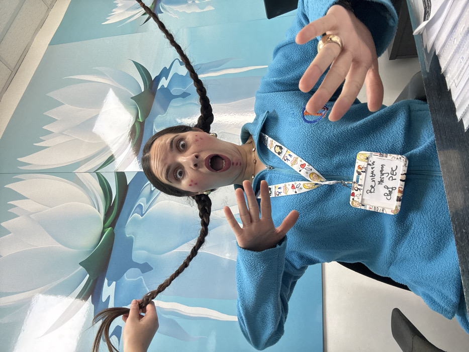 This image captures a woman seated in an indoor office environment, identified by her ID badge as "Benbach Meryam, Chef PEC," located in Meknes, Morocco. The woman, with dark hair styled into two long, braided pigtails extending horizontally from her head, displays a dramatic expression with wide eyes and a widely open mouth. Red markings are visible on her cheeks and around her mouth. She wears a light blue zippered fleece jacket over a beaded necklace. A white lanyard decorated with cartoon characters holds her rectangular ID badge. An unseen individual's hand is visible on the left, grasping and pulling the woman's left braid upwards and leftward. A gold-toned ring is present on the ring finger of her right hand, with both hands raised, fingers splayed. The background features a large wall mural depicting multiple stylized white lotus flowers floating on light blue water with reflections. A dark desk surface is visible in the foreground, supporting a stack of clear plastic pouches or cardholders on the right. A dark office chair is partially visible behind the woman.