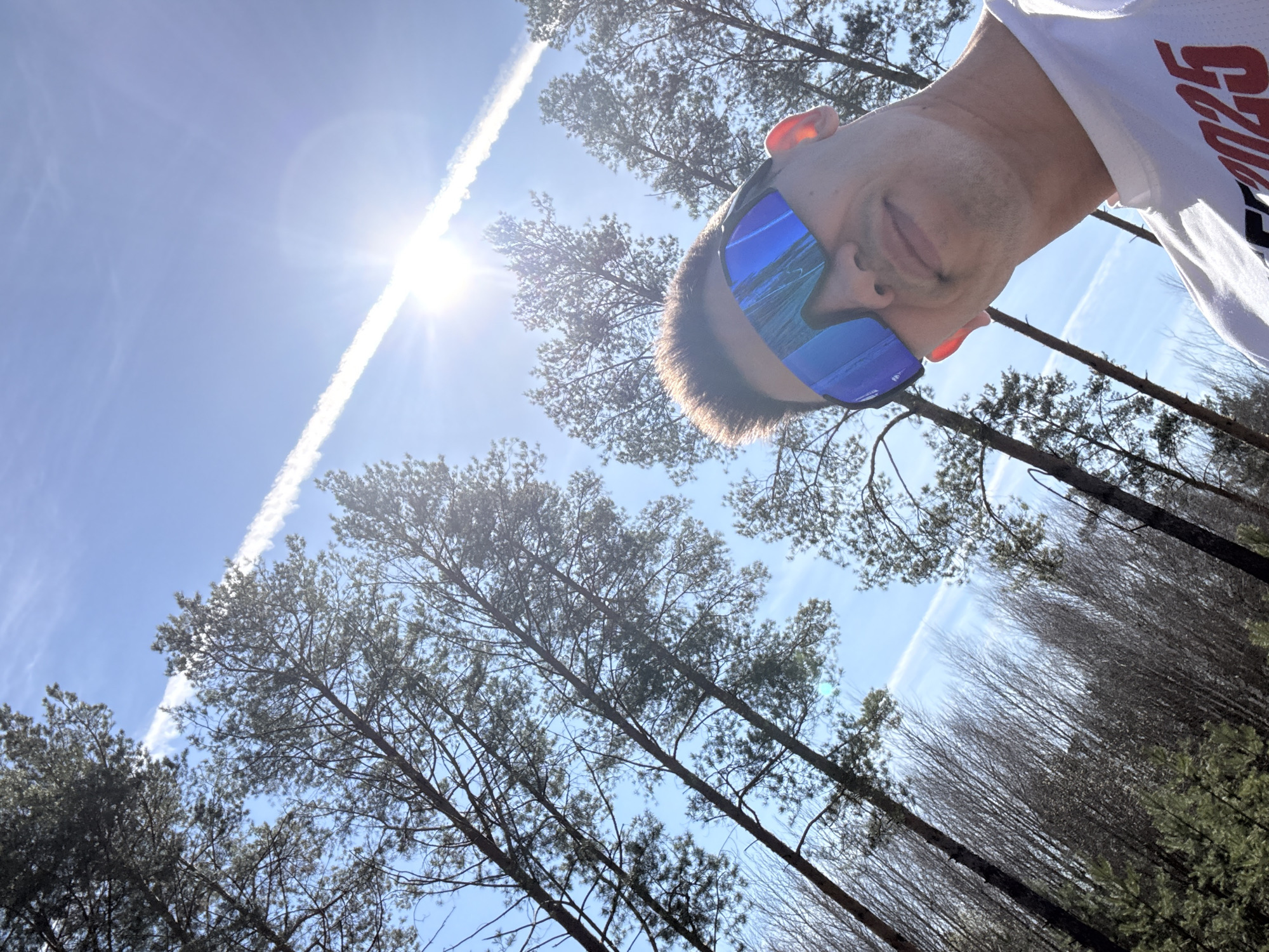A low-angle shot captures a man wearing blue reflective sunglasses and a white t-shirt with red lettering, standing in a wooded area under a bright blue sky. A prominent contrail cuts diagonally across the sky, passing close to the sun. The trees are a mix of bare branches and evergreen foliage. The location is presumed to be a wooded area in Kazan, Russia.