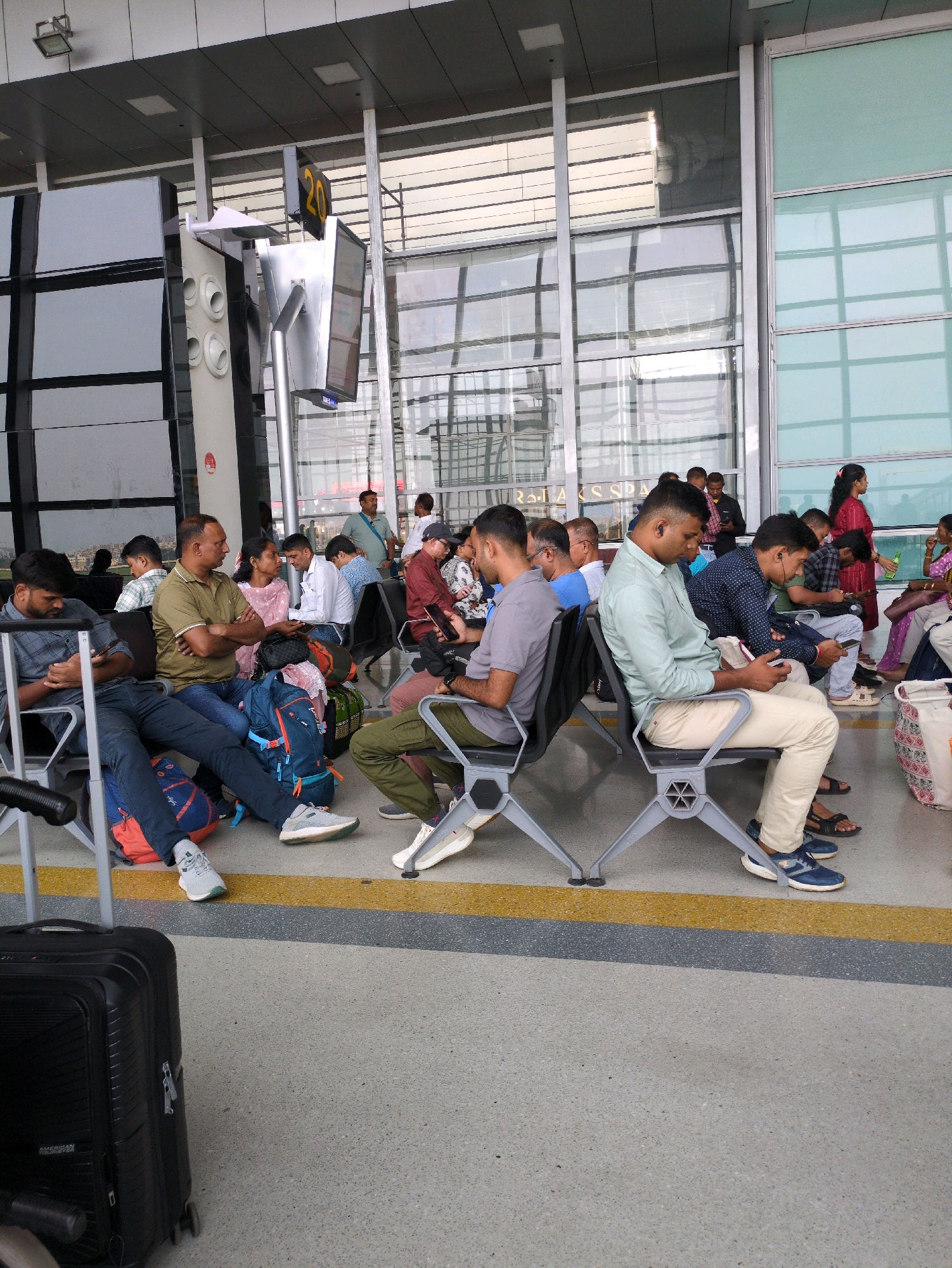 The image depicts a waiting area, likely within an airport terminal, in Dum Dum, India. A group of people are seated on metallic chairs, some looking at their phones, others conversing or resting. Luggage, including backpacks and a rolling suitcase, is visible near the seated individuals. The background features large glass windows, reflecting the interior and exterior of the building, and signage, including a digital display showing the number "20". The floor is paved with concrete and marked with yellow and gray lines. The overall atmosphere suggests a typical scene of travel and transit.