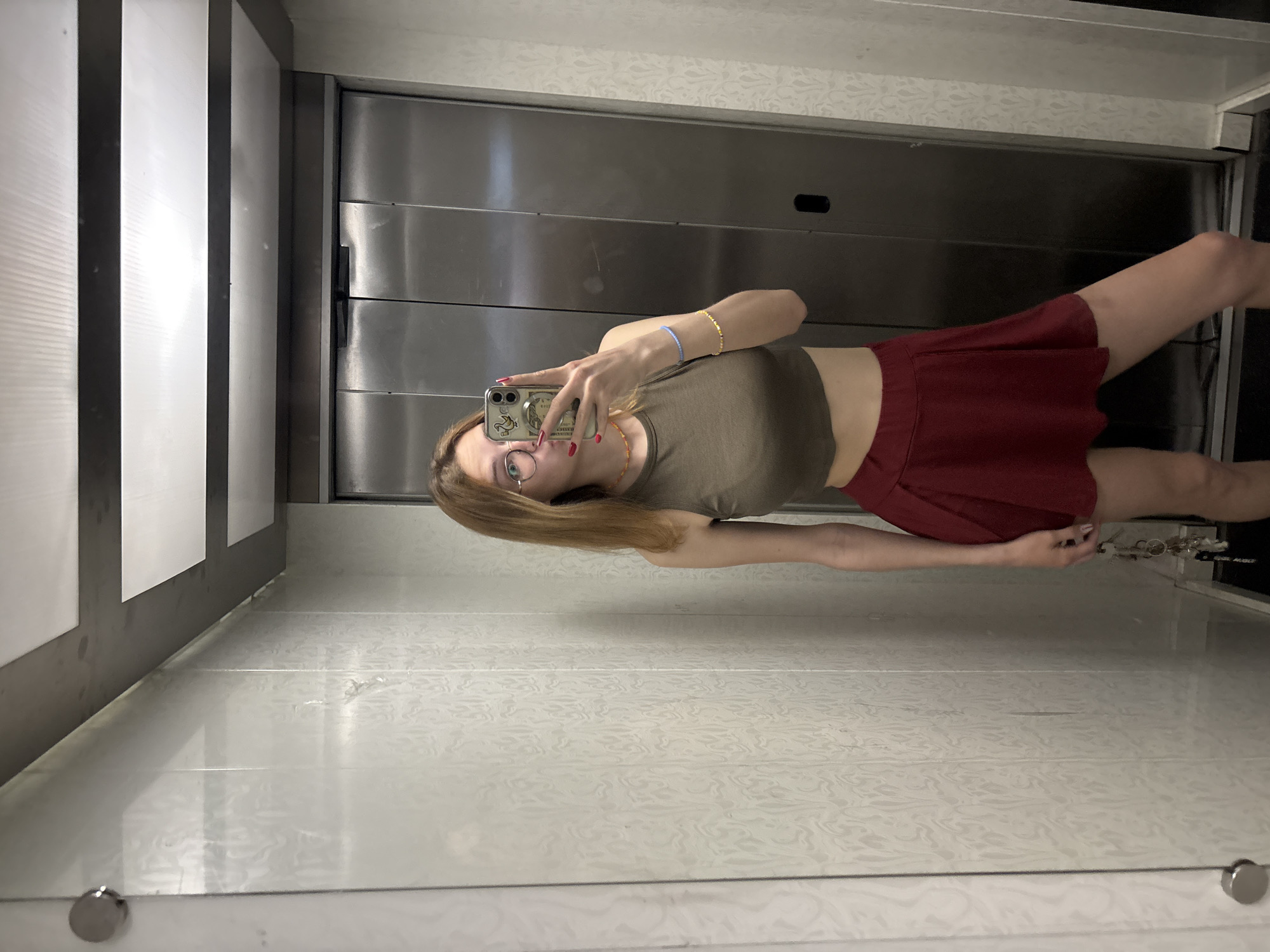 A young woman takes a selfie in an elevator. She's wearing glasses, a brown crop top, and a red skirt. She's holding her phone up in her right hand. The elevator has metallic doors and white patterned walls. The location in Kragujevac, Serbia is not discernable from this image.