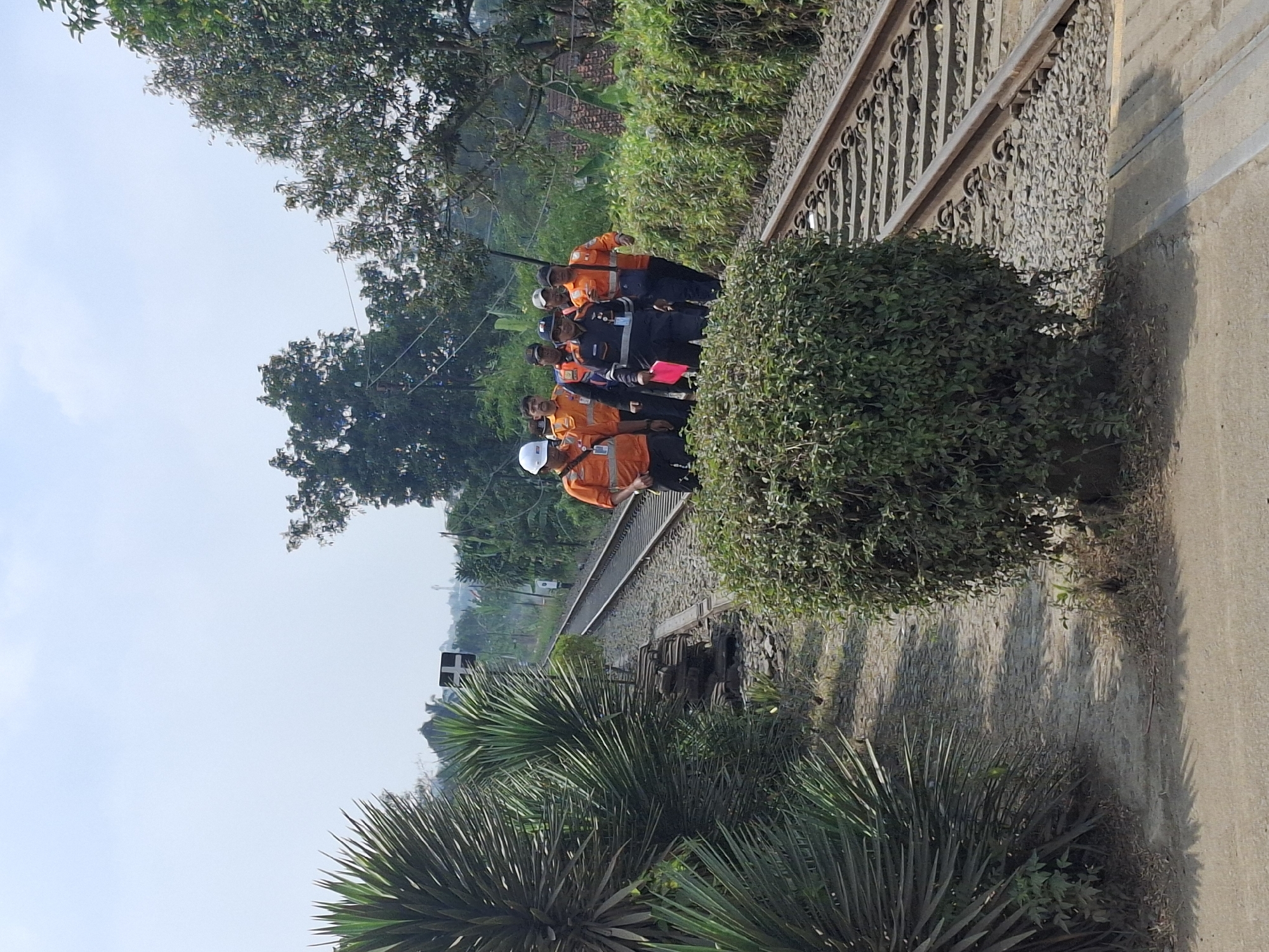 A group of seven individuals, appearing to be workers, walk along a railway track under a clear sky. Five of the individuals are wearing bright orange safety vests over their clothing, and at least two of them have white hard hats. One individual in the front is wearing a dark blue uniform with a badge and is holding a pink folder. The railway track is surrounded by lush green vegetation, including bushes and trees. In the background, there are hints of buildings and structures, suggesting a semi-urban or rural setting. A signal post is visible along the track, indicating the presence of railway infrastructure. The ground around the track is covered with gravel and some weathered wooden sleepers. The overall scene depicts a daylight inspection or survey of the railway line in Ciheulang Tonggoh, Indonesia.