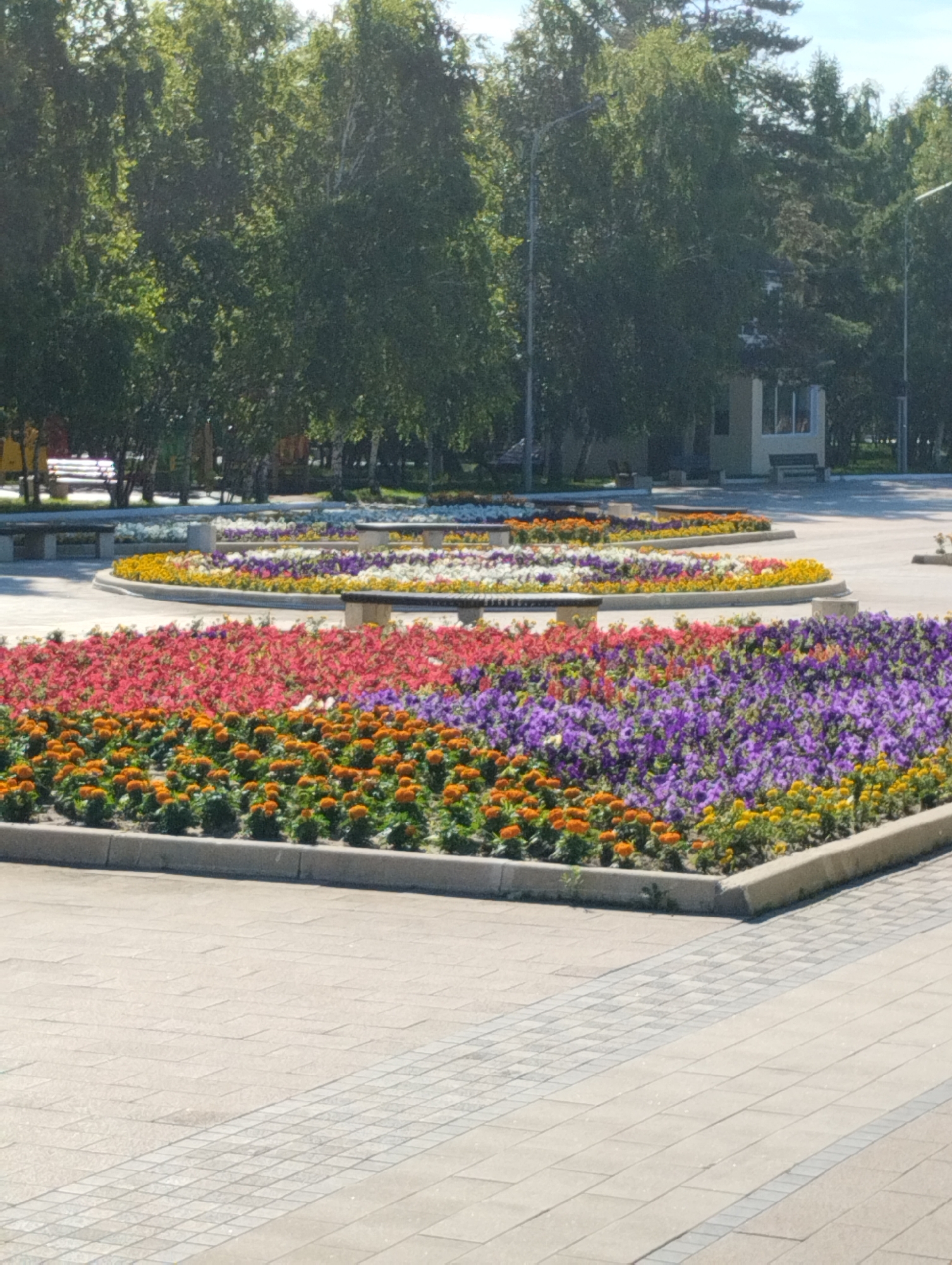 The image shows a park in Kostanay, Kazakhstan, featuring several circular flower beds filled with a variety of colorful flowers. The flower beds are arranged in tiers and planted with a mix of red, pink, orange, yellow, white, and purple blooms. Benches are strategically placed around the flower beds for visitors to rest. In the background, there are trees, a small building, and lampposts, indicating a well-maintained public space. The ground is paved with light-colored tiles, creating a clean and orderly appearance. The scene suggests a pleasant day in the park, with blooming flowers and inviting seating areas.