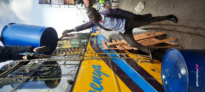 The scene depicts an urban street in Nairobi, Kenya. A large yellow public transport bus, marked with "Kenya" in blue lettering and featuring blue and black stripes, is stationary alongside a paved sidewalk. A metal ladder is affixed to the side of the bus. Two individuals are engaged with a large, blue industrial barrel. One individual, wearing dark trousers and light-colored sneakers, is positioned on the bus's side ladder, handling the blue barrel near the upper section of the bus. A second individual, dressed in a light blue shirt, dark jacket, dark trousers, and dark shoes, stands on the sidewalk below, extending their right arm upwards towards the barrel, appearing to assist in its manipulation. A second identical blue barrel with a white cap rests on the paved ground in the lower left foreground. A disassembled wooden structure lies on the pavement between the bus and the second individual's feet. The background shows blurred elements of a busy street, including market stalls, an advertising board, and distant vehicles and pedestrians, under an overcast sky.