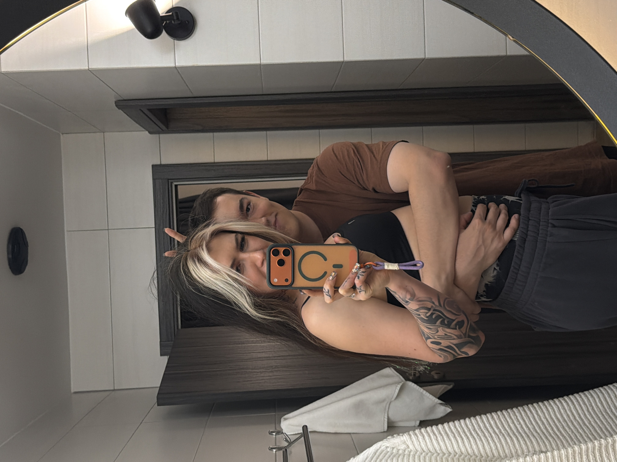 In an indoor mirror selfie, a couple is reflected, capturing an intimate moment. The female subject, positioned closer to the mirror, holds a smartphone in an orange case with a prominent "C-" design and a purple wrist strap to take the picture. She has long dark hair with blonde highlights, a visible arm tattoo, and decorated nails. She is wearing a dark top and dark-colored shorts or pants, and is seen making a hand gesture, possibly a peace sign.
Behind her, the male subject embraces her from behind. He has short dark hair and is wearing a brown t-shirt and dark pants. Both subjects look towards the camera's reflection.
The setting appears to be a modern bathroom or changing area, characterized by white rectangular tiled walls. A dark wooden or laminate counter and shelving unit are visible beneath and behind the couple. Above the mirror, a black light fixture casts a warm glow. Another black circular fixture is seen on the wall to the left. A white folded towel and a white ribbed bathmat or towel are visible in the foreground, below the reflection. The overall scene suggests a casual and affectionate moment, possibly within a private residence or hotel in Palilula Urban Municipality, Serbia.
