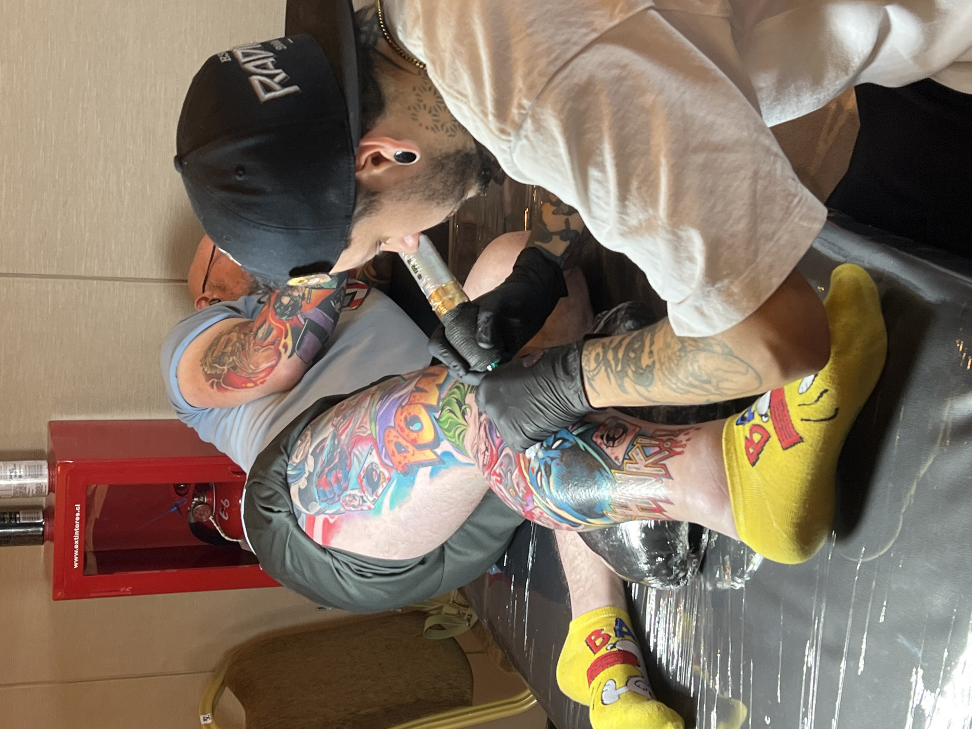 In Arica, Chile, a tattoo artist, wearing black gloves and a black baseball cap, is in the process of tattooing a person's leg. The person receiving the tattoo is wearing yellow socks with cartoon characters. The tattoo appears to be colorful and features comic book characters. In the background, a red fire extinguisher case is mounted on the wall. The scene is set in what appears to be a studio or a similar indoor space.