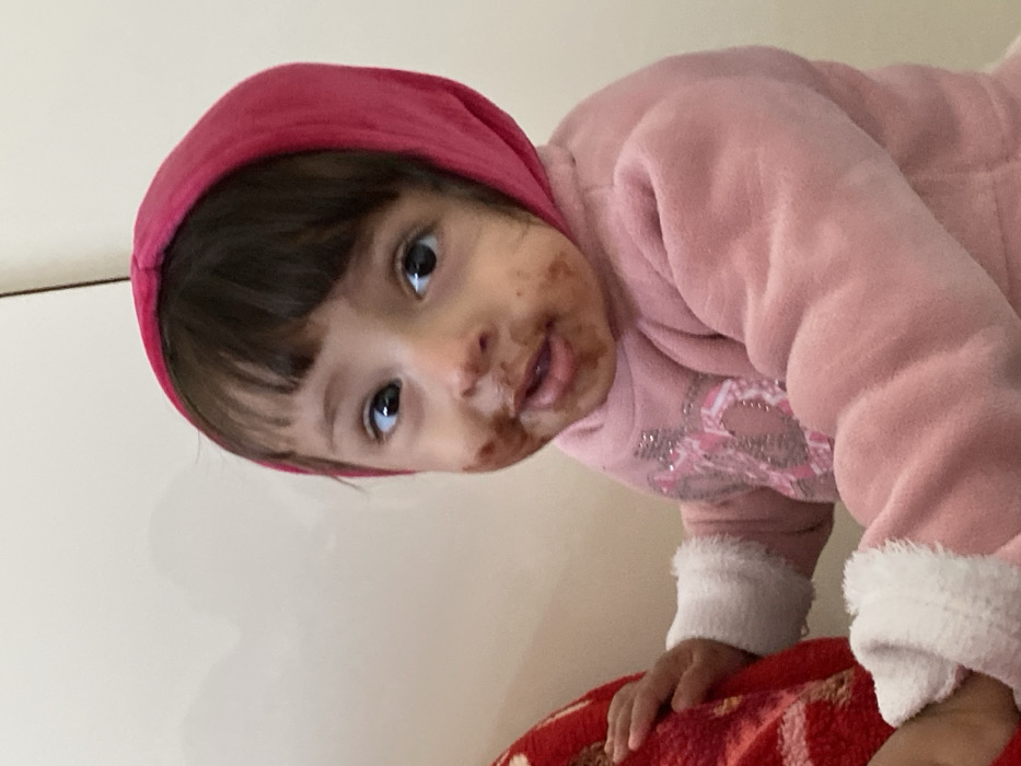 A young child is depicted indoors in Kandahar, Afghanistan. The child, appearing to be a toddler, has dark hair with bangs and dark eyes, looking directly at the viewer. A dark brown, paste-like substance, consistent with food residue such as chocolate, is smeared across the child's mouth, chin, and cheeks. The child wears a pink, soft, possibly fleece-like hooded garment with the hood pulled up, covering the head. The sleeves feature white, fluffy cuffs. Beneath the outer garment, a lighter pink top with a light-colored, possibly silvery graphic design is visible on the chest. The child's left hand grasps a red, textured fabric object, likely a blanket or cushion, which is partially visible on the left side of the frame. The background consists of a plain, light-colored wall with a faint vertical line on the right side.