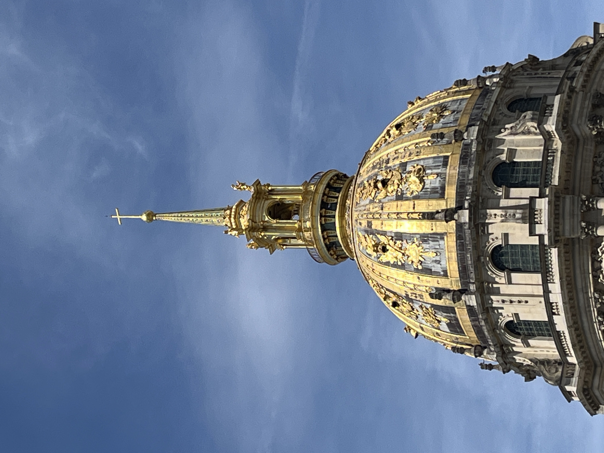 The image shows a close-up view of the golden dome of Les Invalides in Paris, France, against a clear blue sky with sparse clouds. The dome is ornately decorated with gold leaf and features a spire topped with a cross. The architecture is classical, with arched windows and detailed stonework visible on the lower sections of the building. No people or animals are visible in the frame. The lighting suggests it is daytime, likely with bright sunlight given the gleam on the golden dome.