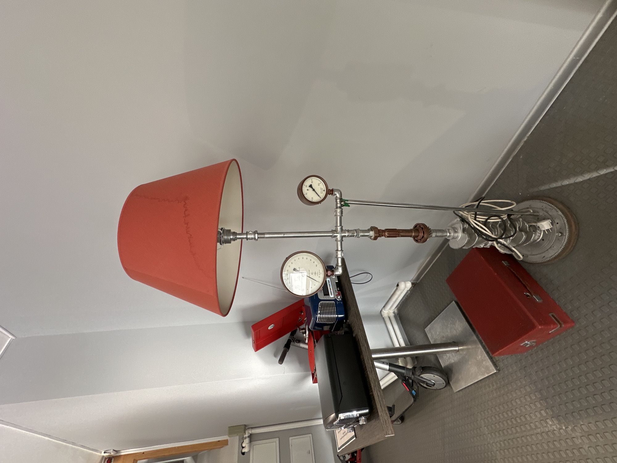 The image shows a red lampshade attached to a lamp made of pipes and gauges. The lamp is positioned on a surface with other objects, including a red toolbox and a black appliance. The background consists of walls and ceilings. There is no information to indicate that the location is in Peresvet, Russia.
