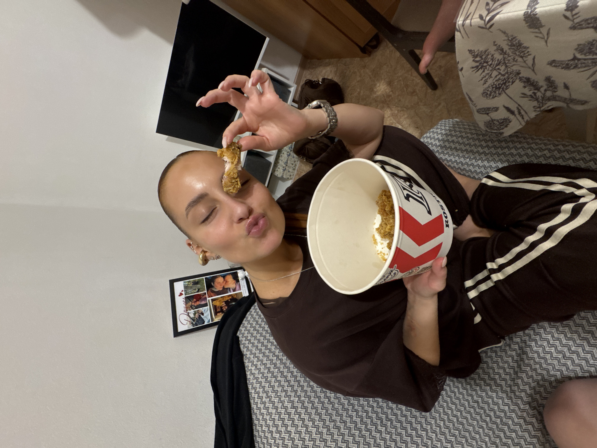 A woman is indoors, holding a piece of fried chicken near her face. She holds a bucket of fried chicken in her other hand. She is wearing a brown top and striped sweatpants, and sitting on a grey patterned surface. The surrounding environment includes a white wall with a framed photo collage, a television, and other furniture. The location is possibly Chaikovsky, Russia.