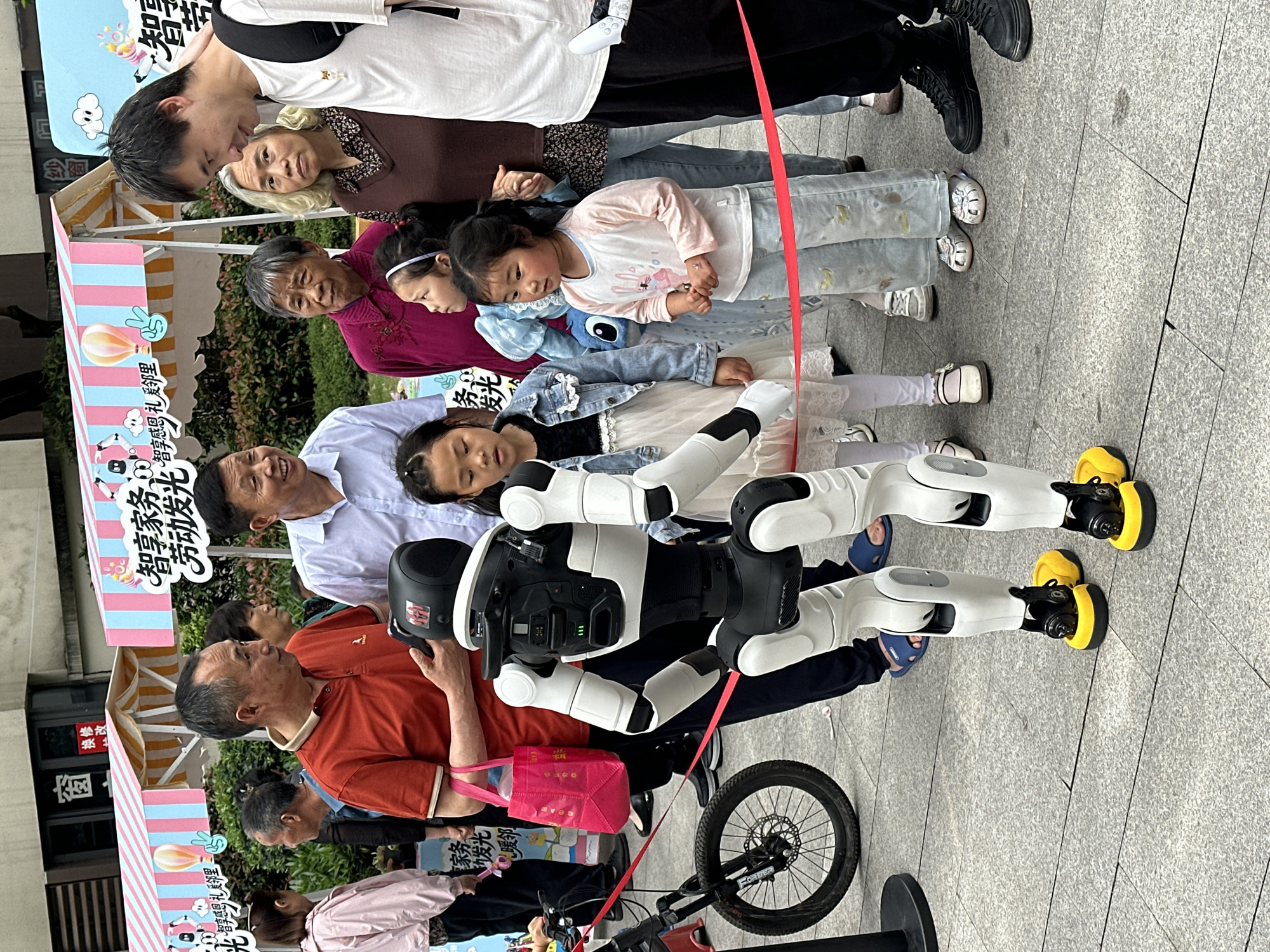 The image shows a diverse group of people, including adults and children, standing in a line next to a black and white robot on a paved surface, which appears to be a sidewalk or public area. A red ribbon is draped across the scene, partially obscuring the robot and people. One person is holding a pink bag. A black bicycle is also present. The background includes signage with Chinese characters, indicating that the photo was taken in the city of Chongqing High-tech Zone, China. The environment appears to be an outdoor public space, potentially during an event or demonstration.