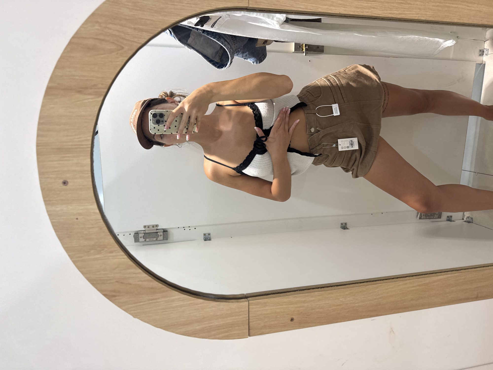 The image shows a young woman in a fitting room. She's wearing a white top with black trim, brown shorts with tags still attached, and a brown baseball cap. She is holding a cell phone with a polka dot case and appears to be taking a selfie in an oval mirror framed in light wood. Behind her, hanging from the top of the fitting room, are jeans and a white shirt. The walls of the fitting room are white. The location and city of Đà Nẵng, Vietnam are not identifiable from the image.