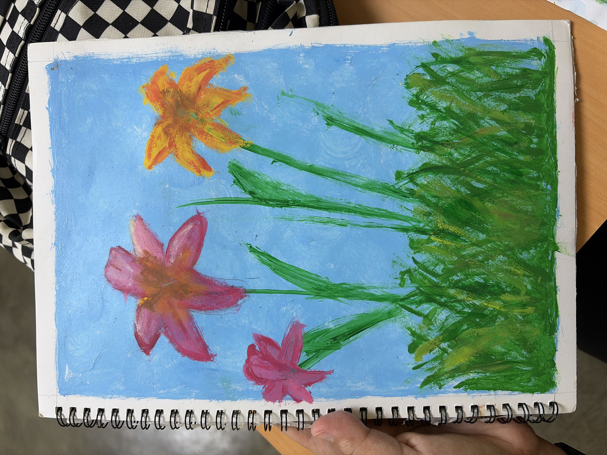 The image is an amateur painting on a spiral-bound sketchbook. The painting depicts three flowers with green stems against a blue background. One flower at the top is orange, while the other two are pink. To the right of the flowers is a dense patch of green brushstrokes representing foliage. The sketchbook is resting on a wooden surface and partially held by a human hand. The background includes a checkered backpack. There is no indication that this image shows Guayana City, Venezuela.