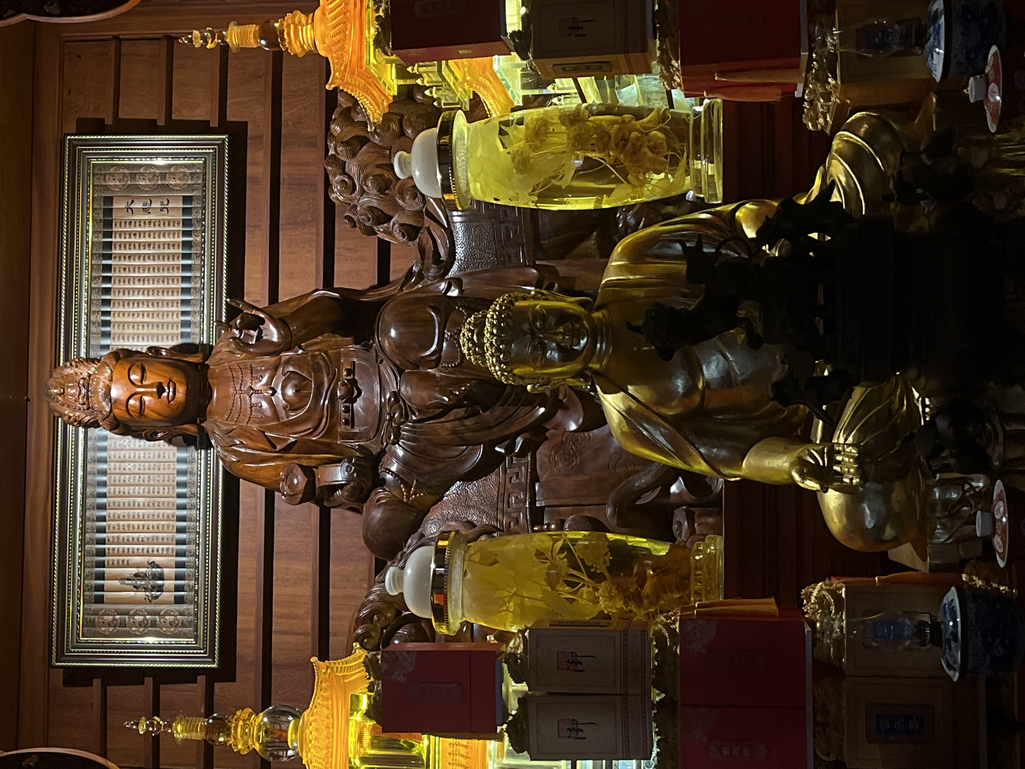 The image depicts a shrine within a temple or religious establishment in Mũi Né, Vietnam. The scene is dominated by two Buddha statues, one carved from wood and positioned higher up, seated on what appears to be a lion or mythical creature. This wooden Buddha is adorned with a crown and has its right hand raised in a gesture. Below it, a second, larger Buddha statue is made of a golden, metallic material, seated cross-legged with its hands resting in its lap. The backdrop is a wooden paneled wall, with a framed artwork featuring scrolls and what seem to be depictions of Buddhas or religious figures. Flanking the statues are tall, decorative glass jars filled with a yellow liquid and submerged botanical specimens, possibly medicinal herbs or roots. To the sides, there are illuminated structures resembling pagodas or architectural elements, glowing with orange and yellow light. Red boxes with gold Chinese characters are also visible, stacked and arranged around the display. In the foreground, below the golden Buddha, a dark, intricately carved incense burner or censer is visible. The overall atmosphere is one of reverence and tranquility, illuminated by warm, ambient lighting.