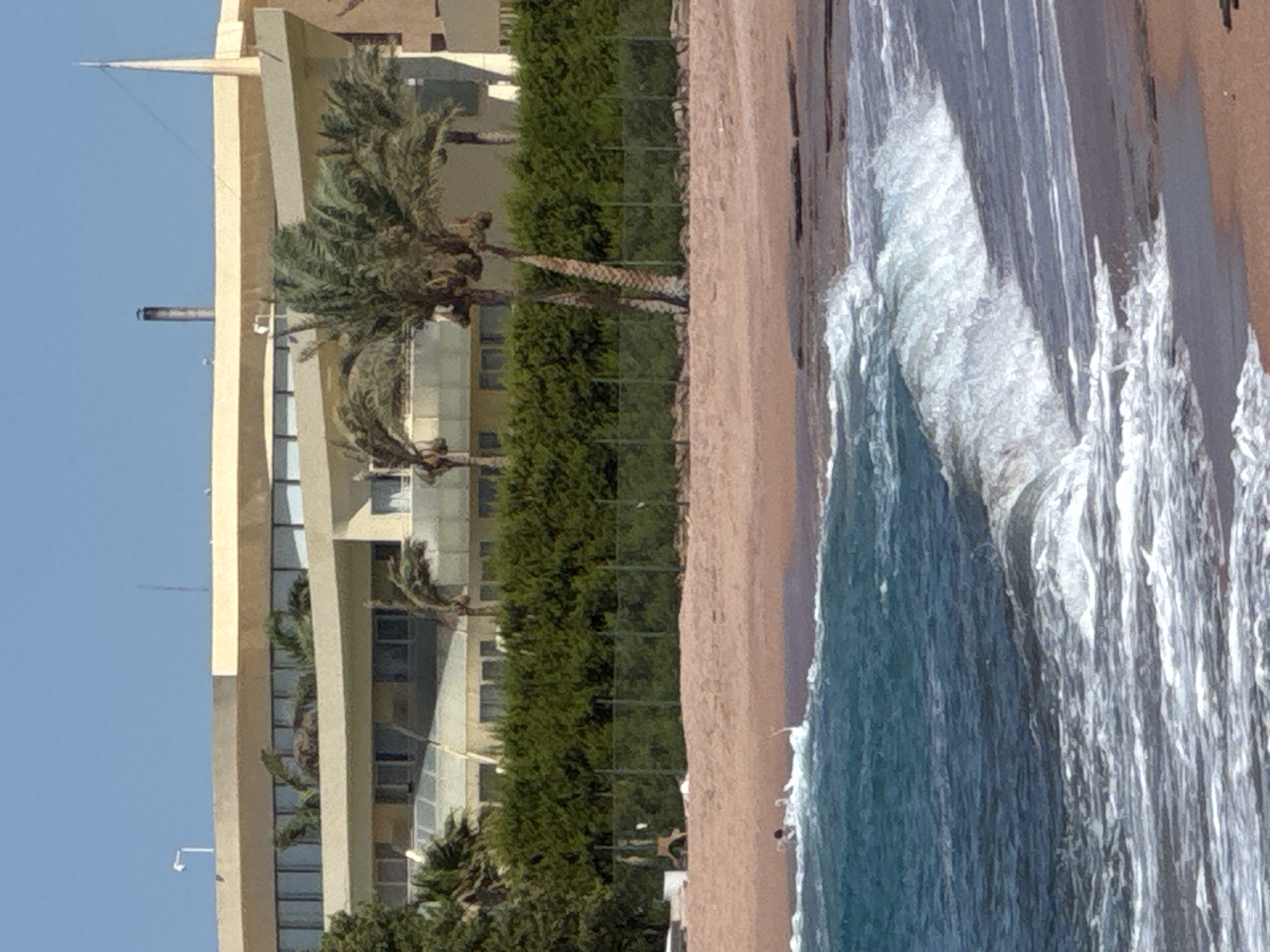 The image depicts a beach scene with a multi-story building and palm trees in the background. The building is light-colored with a curvilinear design and numerous windows. Lush green foliage borders the sand, and a fence runs along it. In the foreground, the ocean is visible with waves crashing on the shore. A few individuals are in the shallow water, appearing to be swimming or wading. The sand on the beach is a light brown color. The sky is clear and blue.