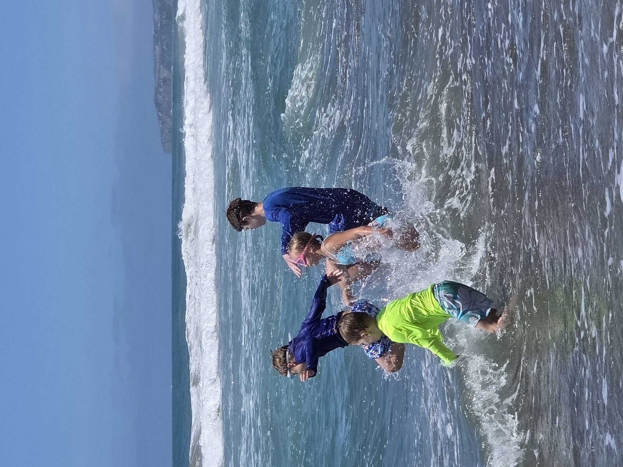 This is a full-frame, eye-level, outdoor, vertical, natural light photograph of four children playing in the ocean waves at a beach. The two older children, a boy and a girl, are on the right side of the frame. The boy, wearing a blue long-sleeved rash guard, is reaching out and splashing. The girl, wearing a blue bikini and pink goggles, is in the center, also splashing. To the left, a younger boy in a blue long-sleeved rash guard and patterned swim trunks is jumping or lunging forward, with goggles on. In the foreground on the left, a young boy in a bright yellow long-sleeved rash guard and patterned swim trunks is visible, also playing in the waves. The ocean is a deep blue-green with foamy white waves breaking. In the background, the sky is a clear blue with some hazy clouds. A distant cliff face, possibly representing the coastline of San Clemente, California, is visible on the horizon. The overall scene captures a moment of joyful activity and play in the ocean.