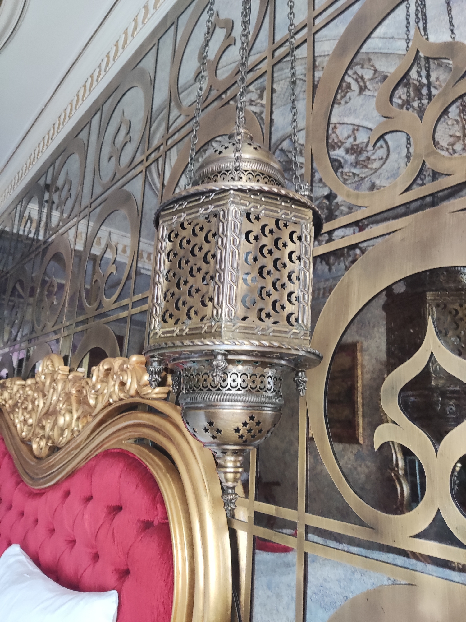 The image displays a close-up of an ornate, metallic lantern hanging from a chain. The lantern is intricately designed with cutouts in the shape of crescent moons and stars, suggesting an Islamic or Ottoman influence. It is positioned in front of a decorative metal screen with elaborate scrollwork and geometric patterns. To the left of the lantern and screen is a plush, red upholstered headboard of a bed, adorned with gold trim and tufting. A white pillow is partially visible on the bed. The background appears to be a wall with decorative panels and a mirrored surface, reflecting parts of the room. The overall aesthetic suggests a luxurious and historically rich interior, possibly within a hotel or a meticulously decorated residential space in Beyoğlu, Turkey.