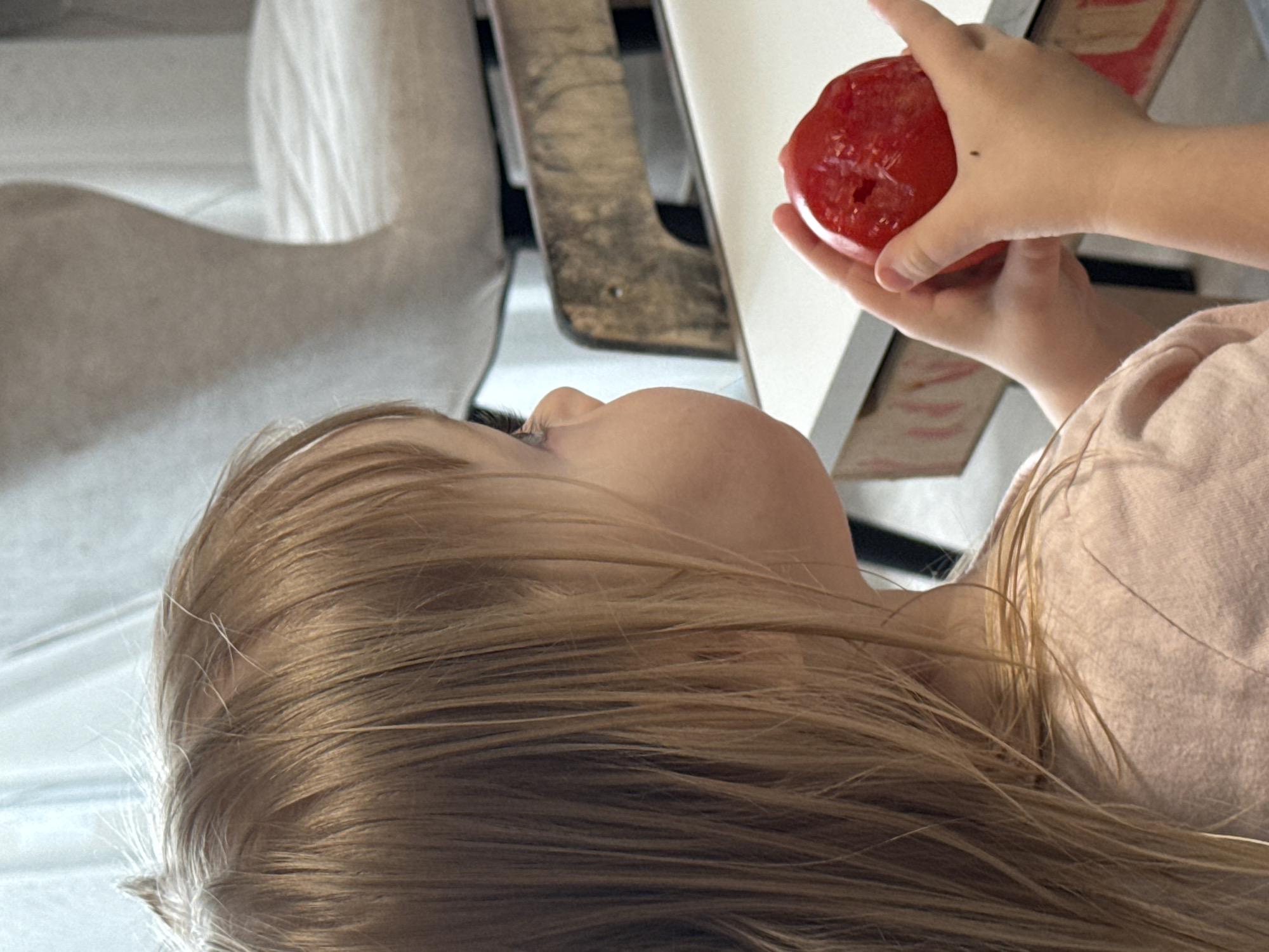 A young child with blonde hair looks up at a red object held in another child’s hands. The background includes a white chair and a wooden chair. The location and city are unknown.