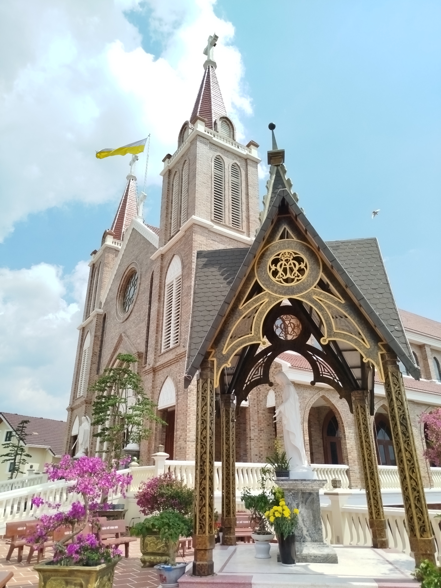 The image captures the exterior of a church, possibly a Catholic church, in Cam Ly - Đà Lạt, Vietnam. The building is constructed of light-colored brick, featuring a tall, central spire topped with a cross, and a yellow and white flag fluttering from one of the secondary towers. The architectural style includes arched windows and decorative elements.
In the foreground, a smaller, ornate structure with a peaked roof and decorative metalwork houses a white statue, likely of the Virgin Mary. The area surrounding the statue is adorned with potted plants and flowers, adding color to the scene. The foreground also includes stone benches and a short flight of stairs leading up to the building. The sky is blue with scattered clouds, indicating a clear, sunny day. A bird is flying in the sky on the right side of the statue canopy.