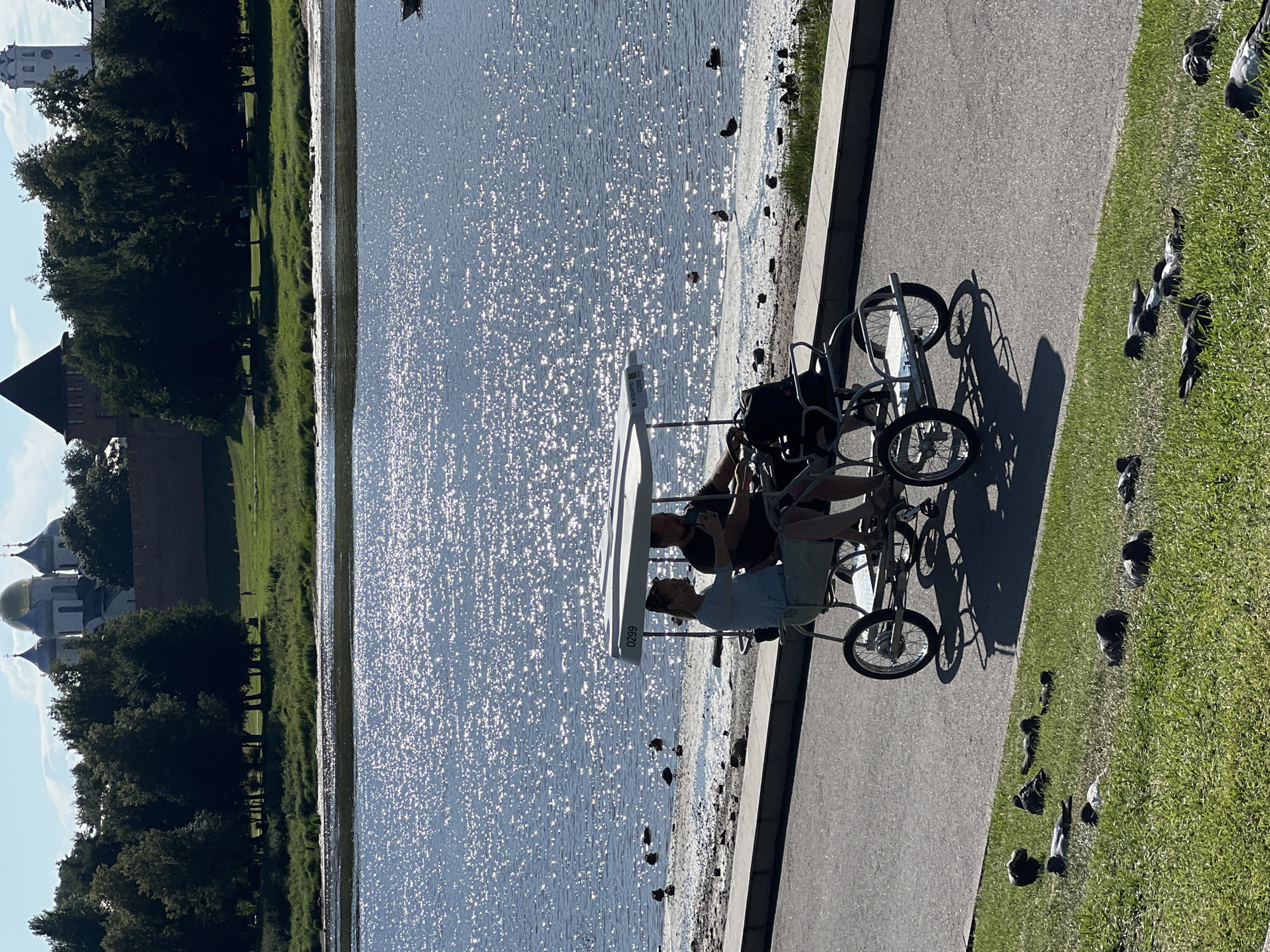 In Veliky Novgorod, Russia, on a sun-drenched day, a tandem quadricycle carrying two individuals is parked on a paved pathway beside a body of shimmering water. The couple, a man and a woman, are seated in the quadricycle. The woman, on the left, wears a light blue top and dark pants, while the man, on the right, is dressed in a dark t-shirt and black shorts. The man is holding a mobile phone. In the background, across the water, a serene landscape unfolds with a line of trees, a grassy embankment, and a red brick wall, beyond which distinctive architectural structures with golden domes and spires rise against a clear blue sky, characteristic of historic religious buildings in Veliky Novgorod. Numerous ducks are visible on the gravelly shore of the water. On the grassy area in the foreground, several pigeons are resting or pecking at the ground. The overall scene depicts a leisurely moment in a scenic historical setting.