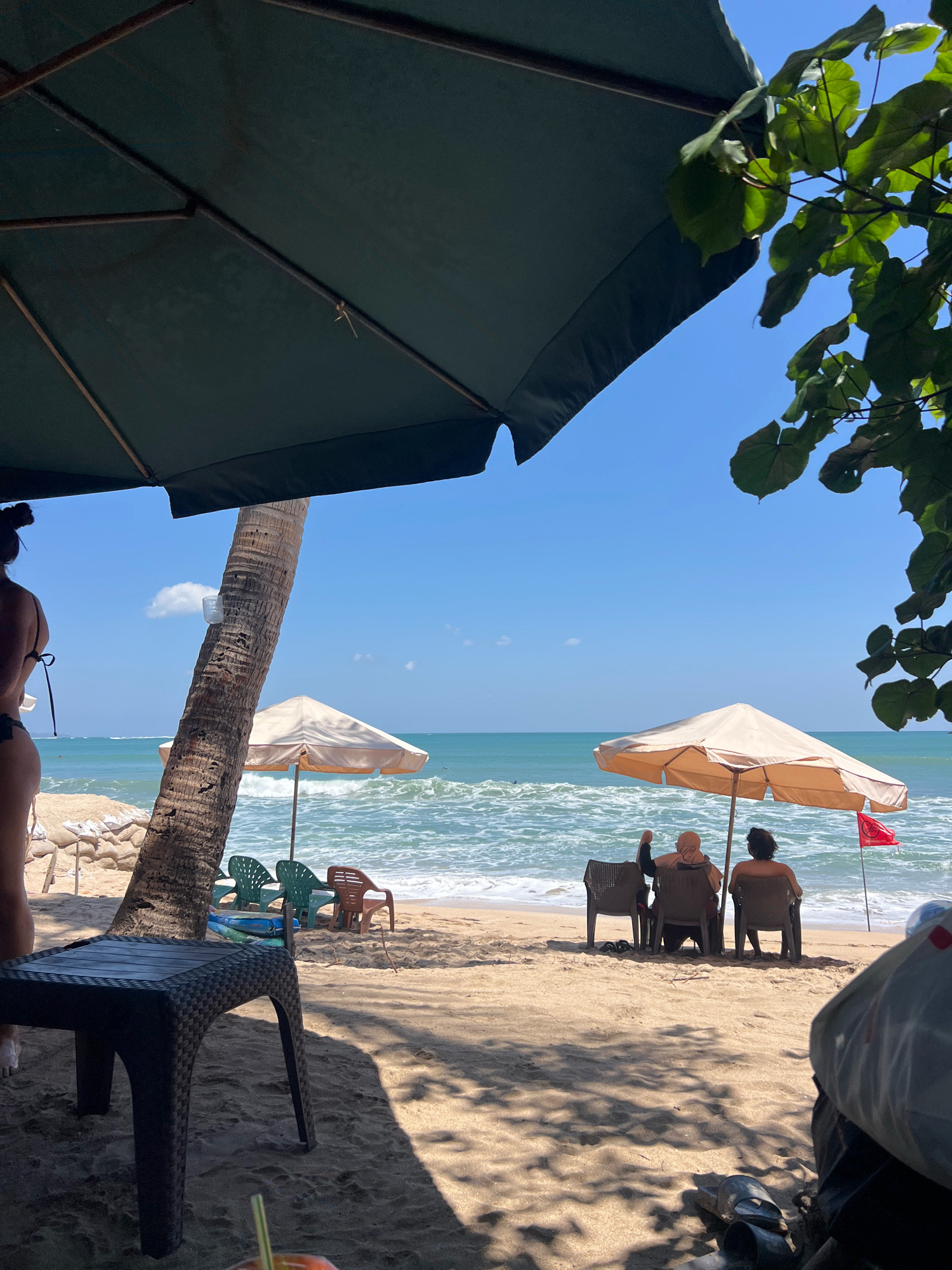 In Kuta, Indonesia, on a sandy beach under a clear blue sky, a scene unfolds near the ocean. Two people sit in chairs beneath a large, light-colored umbrella, facing the water. A red flag flies nearby on a thin pole. To their left, another umbrella, this one dark green, covers a table and part of a palm tree trunk. In the foreground, a dark plastic table is positioned on the sand, with a shadowy area cast by the umbrella. Further back, several empty green chairs are visible, along with a brown chair. The ocean has moderate waves, with white foam breaking on the shore. A person in a dark bikini is partially visible on the left side of the frame, facing away from the camera. The overall impression is of a relaxed day at a beach resort.
