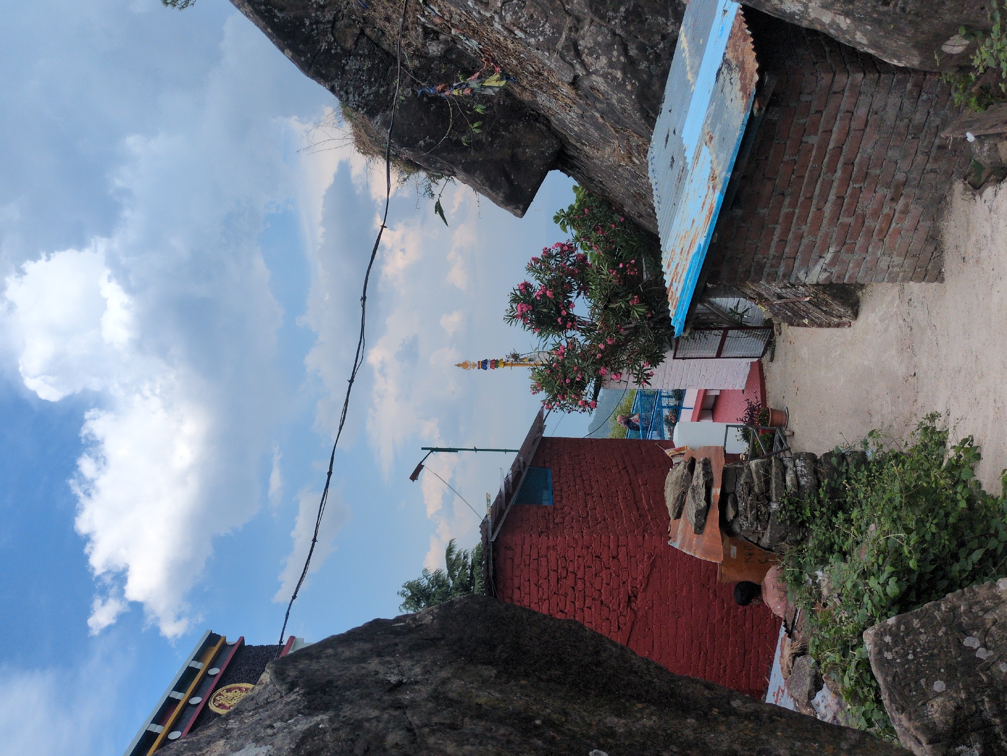 This image depicts a mountainous area in Doh, India, with a narrow pathway leading between large rock formations and structures. On the left, a red brick building with a blue-trimmed roof is visible, next to a rough stone wall. A person is seated near this building. On the right, another structure with a corrugated metal roof, also with blue trim, is built against a rocky outcrop. This outcrop features prayer flags attached to it. In the background, a stupa-like structure with colorful banners rises above a blooming pink-flowered bush. The sky is partly cloudy.