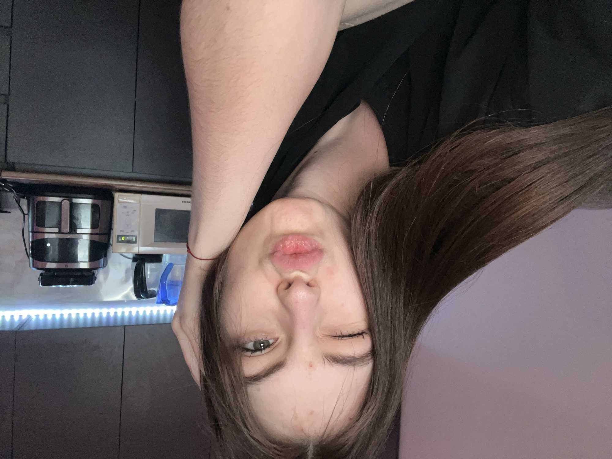 A young woman is posing indoors. She has fair skin, brown hair, and is wearing a black shirt. She is winking one eye and pursing her lips. Her arm is raised and bent near her head. In the background, kitchen appliances are visible on a countertop with LED strip lighting underneath. Cabinets are behind the appliances. A pink wall is visible to the side. The location cannot be determined to be in the city of Saint Petersburg, Russia.