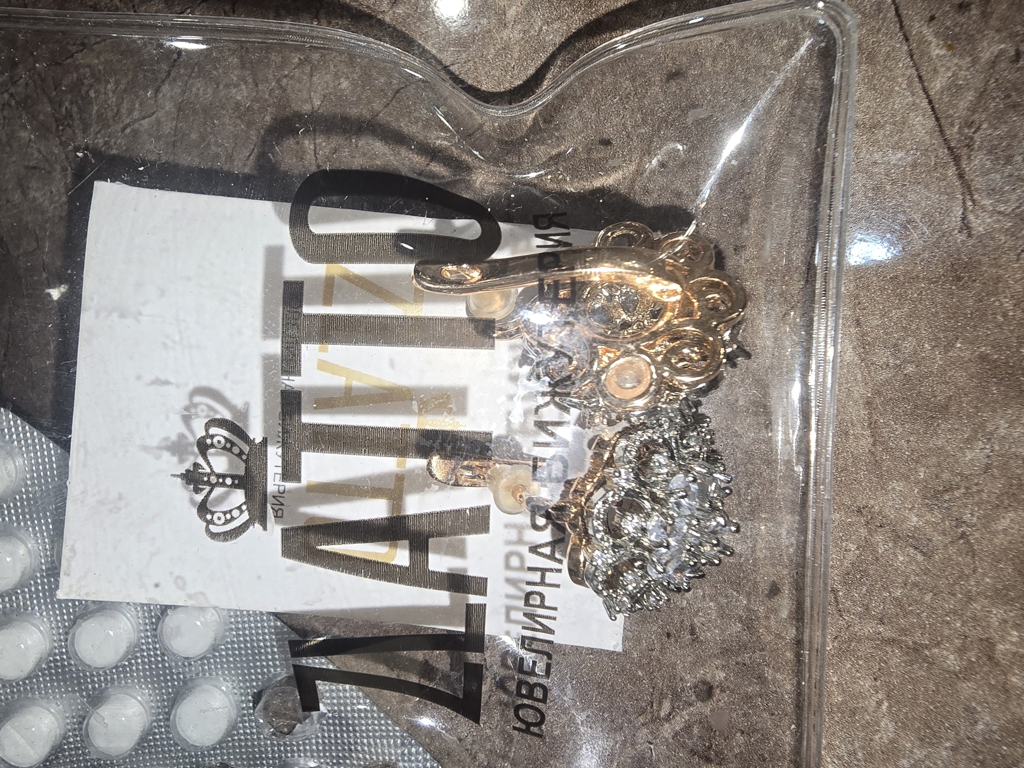 The image shows a close-up of a clear plastic package containing a pair of gold-colored earrings with clear stones. The earrings are ornate, with one appearing to be a stud and the other a dangle style. Behind the earrings, a white card with a black crown logo and the word "ZLATTO" in large, stylized letters is visible. Below "ZLATTO", the text "Ювелирная бижутерия" (Jewelry costume jewelry) is printed in Russian. The background is a textured, brown surface, possibly a table or countertop. A blister pack containing white pills is partially visible in the upper left corner. The image does not provide any information about the location in the city of Ulan-Ude, Russia.