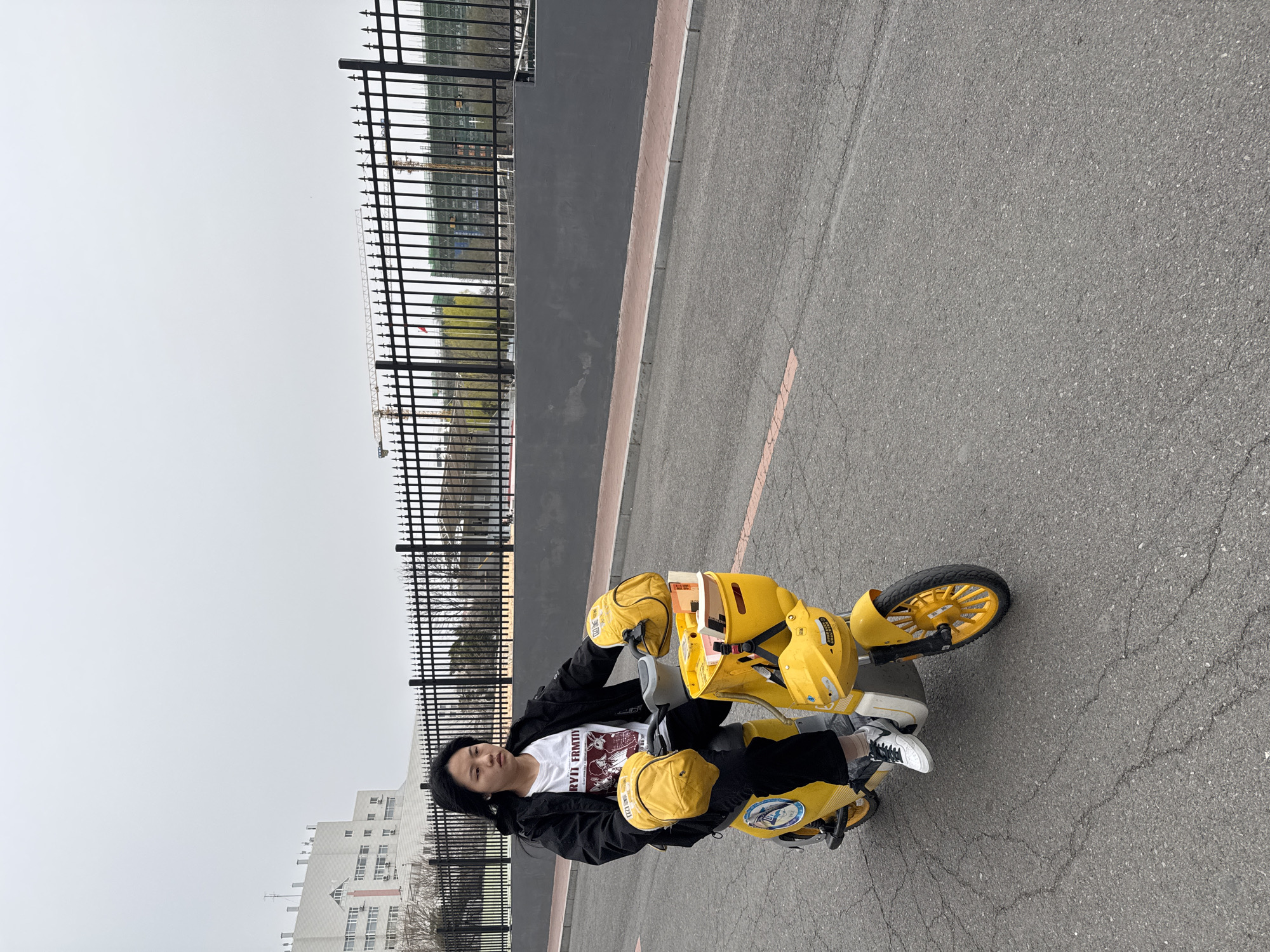 A person is sitting on a yellow bicycle in Zhongtun, China. The person is wearing a white t-shirt, black pants, and white sneakers. The bicycle is parked on an asphalt road. Behind the person and the bicycle, there is a black metal fence and a grey wall. Beyond the fence, buildings and overcast sky are visible.