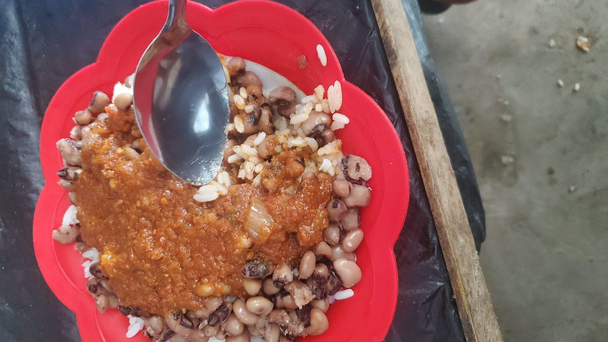 A close-up, overhead shot shows a red, scalloped plastic plate filled with a meal. The plate is placed on a dark, possibly black, plastic surface, which is itself resting on a rough, wooden plank. The meal consists of white rice topped with black-eyed peas and a reddish-brown stew. A silver spoon rests on the plate, partially submerged in the food, with its handle extending out of frame to the right. The background is dark and out of focus. The overall setting suggests a casual dining environment, likely outdoors or in a simple eatery. The lighting is dim, with reflections visible on the spoon. The image composition focuses on the food and plate, with the surrounding environment less defined.