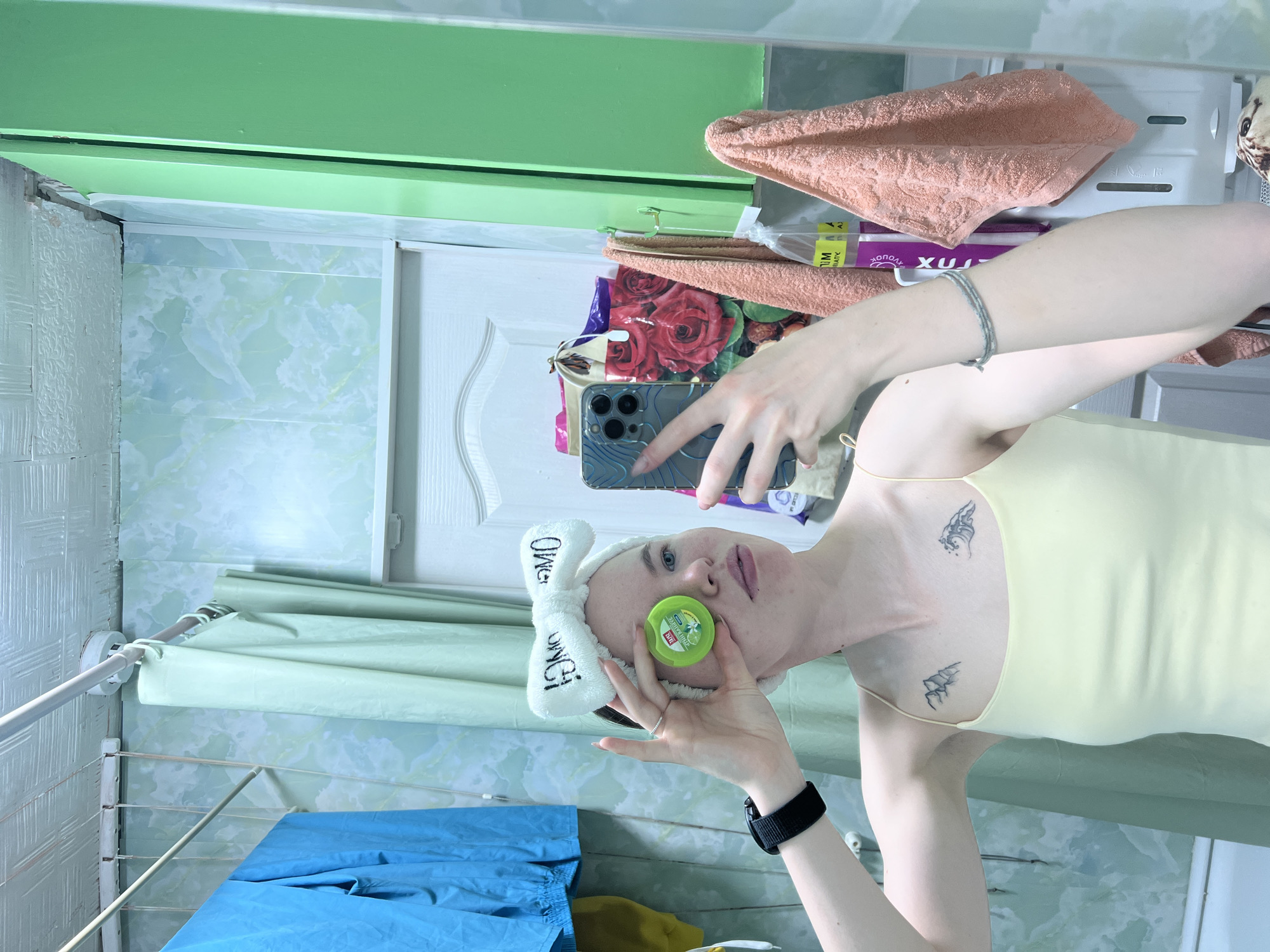 A woman is taking a selfie in what appears to be a bathroom or washroom. She is wearing a white headband with the words "OMG! OMG!" written on it, a light yellow tank top, and a black smartwatch. She is holding a green circular object against her face, possibly a beauty product or medicated balm. Her phone is in her other hand, pointed at her own face. There are tattoos visible on her chest and shoulders.
The background shows a tiled wall, a drying rack with blue and green clothing, and a bathroom cabinet with towels and other items. The style and details of the bathroom suggest a residential setting.
The image has aspects consistent with an interior scene in a home in Chaikovsky, Russia, based on the architectural style and design of the bathroom. However, additional contextual information would be required to definitively confirm the location within that city.