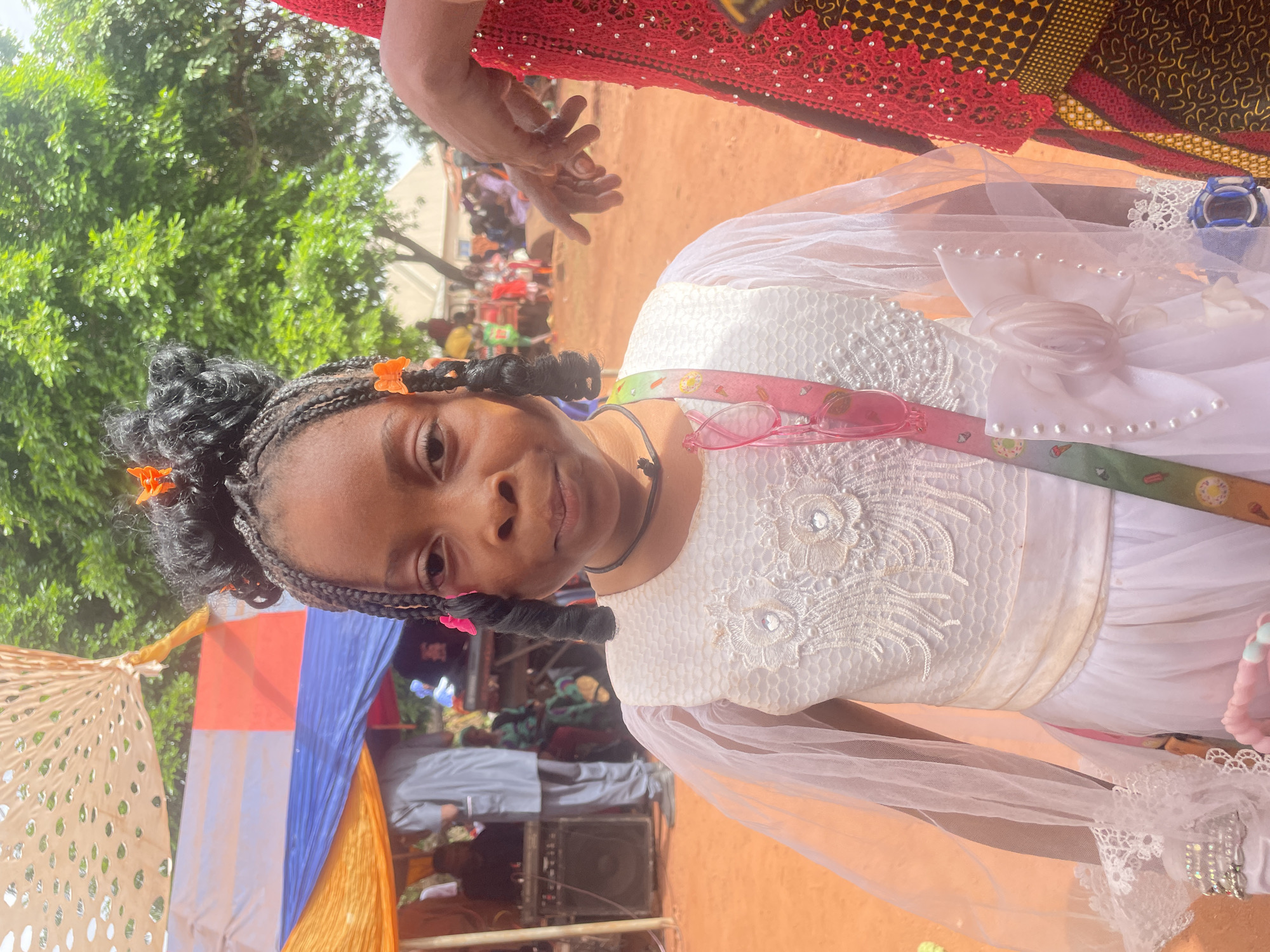 A young girl in a white dress stands outdoors in what appears to be a public gathering in Ogbe, Nigeria. The dress features lace detailing and sheer sleeves. She has braided hair with small orange and pink flower clips. She wears a ribbon sash with colorful patterns. In the background, there are people, trees, and tents. One person wears red and yellow patterned cloth. A speaker is visible near the tents. The ground is a reddish-brown color.