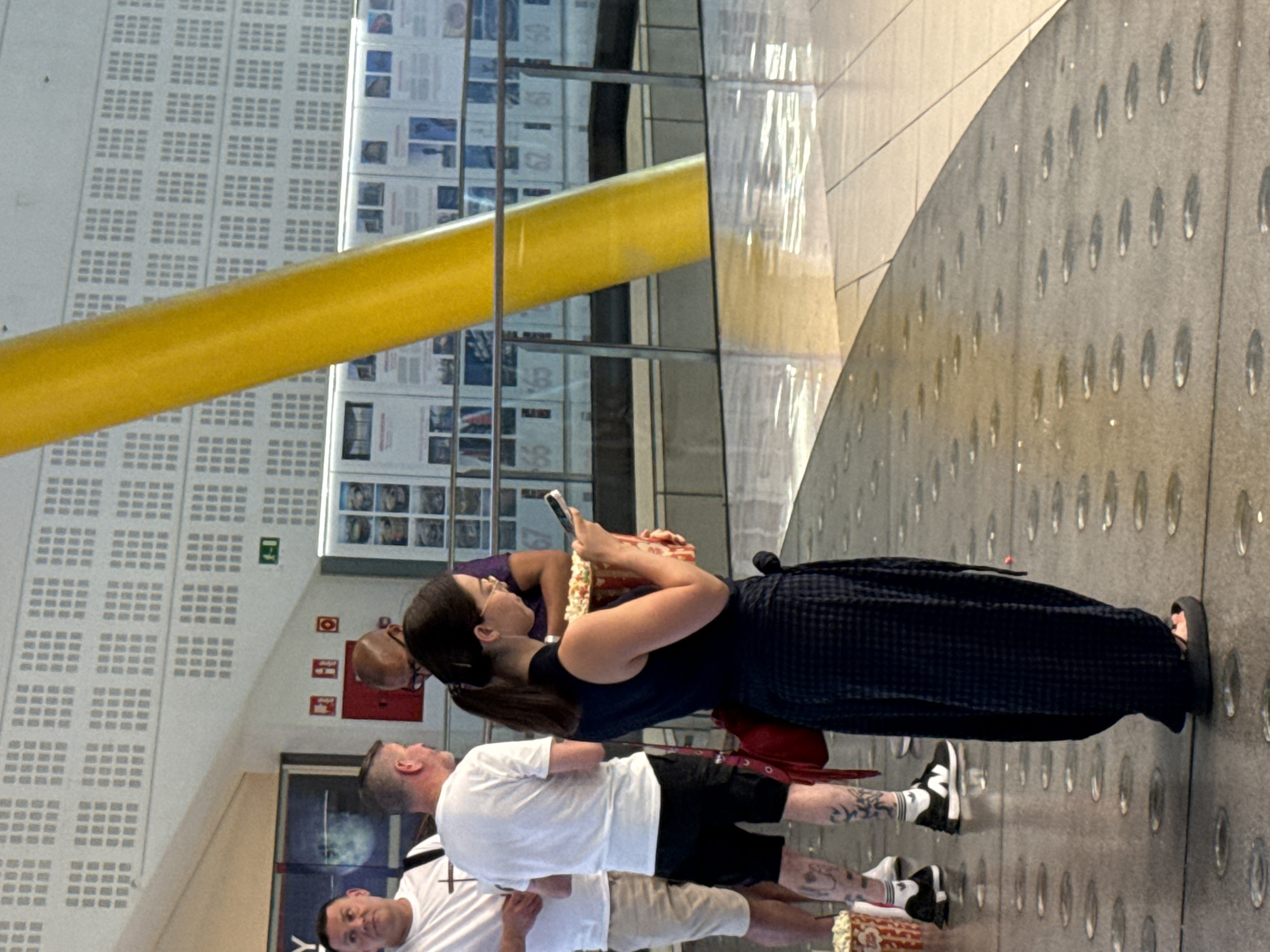 The image shows a group of people inside a modern building, possibly a museum or entertainment venue, with large informational displays on the wall in the background. A bright yellow column is a prominent feature. A woman in the foreground is holding a large container of popcorn and looking at her phone. She is wearing a dark sleeveless top and a dark patterned skirt. Two men are standing behind her; one is wearing a white t-shirt and tan shorts, and the other is wearing a white t-shirt and black shorts with tattoos on his legs. The flooring is a polished surface with circular indentations, reflecting the light. The architectural style and the presence of informational displays suggest an indoor public space. The image quality is somewhat blurry, indicating it was taken with a mobile device.