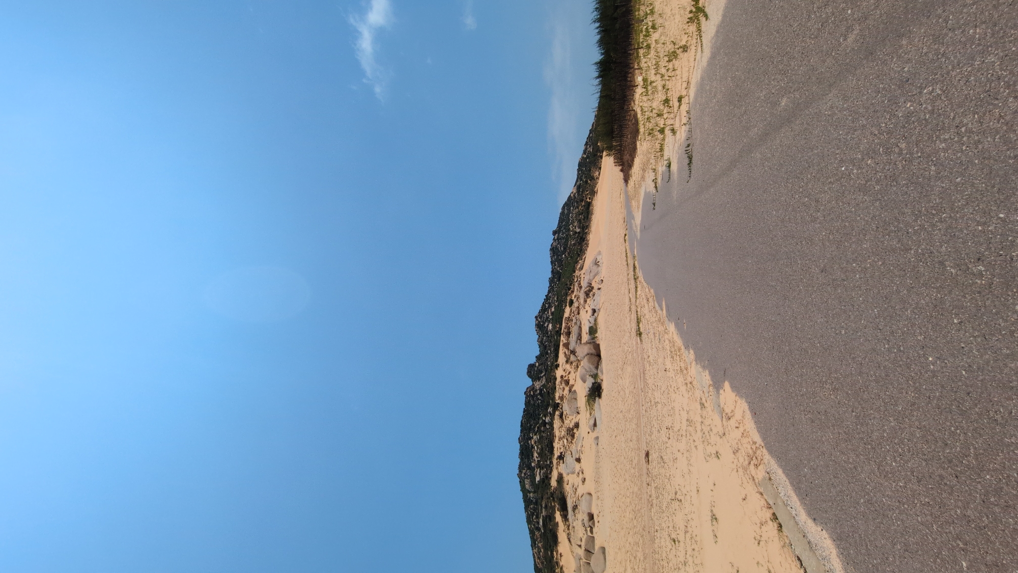 The image depicts a landscape in Phuoc Dinh, Vietnam. A paved road runs along the right side of the image. To the left of the road, there's sandy terrain with sparse vegetation and rock formations. A line of trees is visible in the upper right corner. The sky is clear blue with a few light clouds.