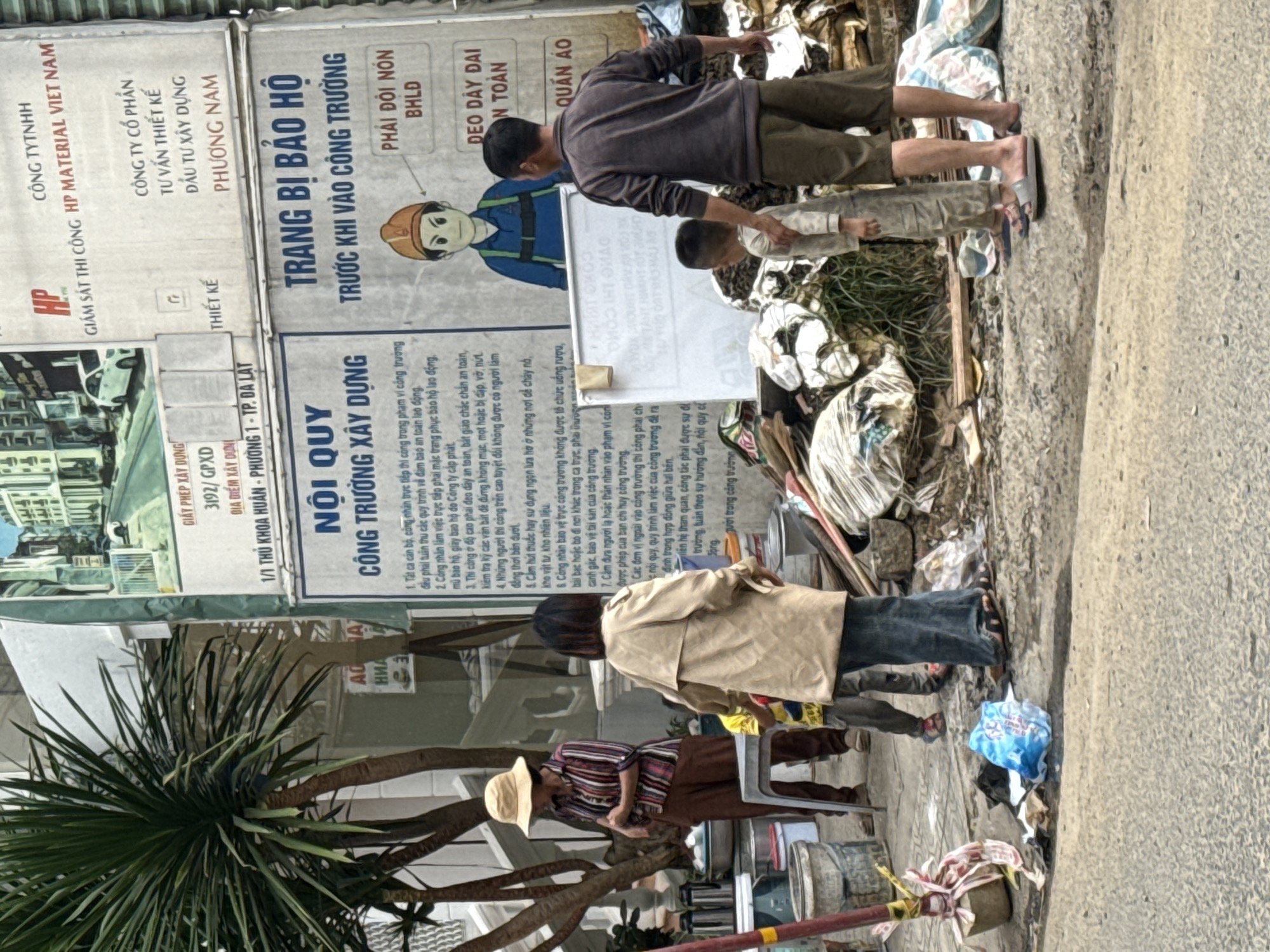 A group of people, including adults and children, are standing on a sidewalk next to a construction site. The sidewalk is littered with trash, including plastic bags, debris, and construction materials. In the background, there is a building with various signs, including one that reads "NỘI QUY CÔNG TRƯỜNG XÂY DỰNG" (Construction Site Regulations). Another sign advertises "TRANG BỊ BẢO HỘ TRƯỚC KHI VÀO CÔNG TRƯỜNG" (Safety Equipment Before Entering the Construction Site). A cartoon drawing of a construction worker is displayed on this sign, with instructions on what safety gear to wear, such as a helmet and protective clothing. One adult woman, wearing a hat and a striped shirt, stands to the left, looking down. Another adult woman, wearing a light-colored coat, stands in the center, facing away from the camera. Two children are present, one boy in a light-colored shirt and pants, and another younger boy or girl in a yellow shirt. An adult man, wearing a dark shirt and shorts, stands to the right, with his arm around the younger child. The environment suggests a gritty urban setting in Da Lat, Vietnam, with a mix of construction activity and accumulated waste.