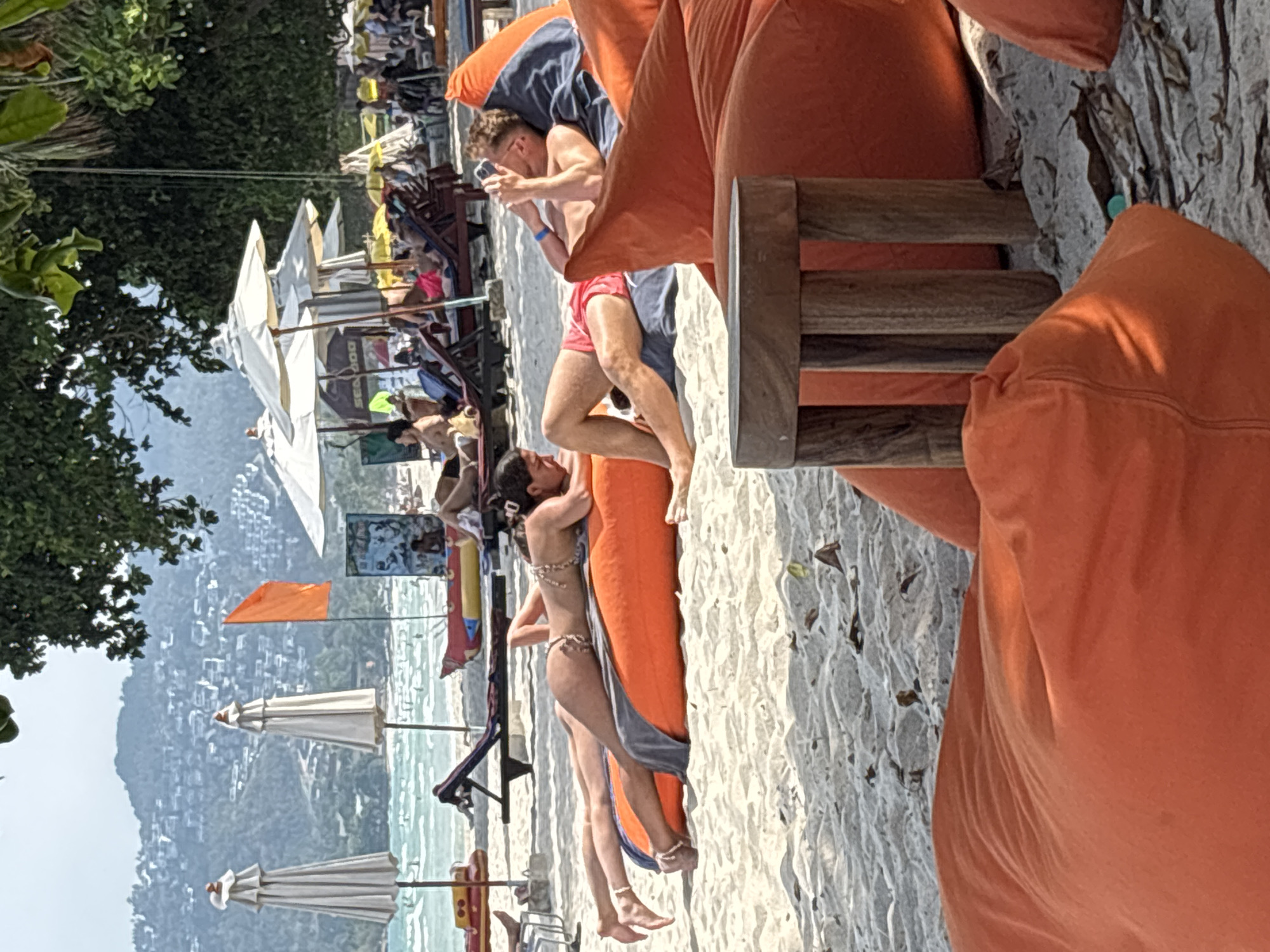 The image depicts a vibrant tropical beach scene in Baan Chaweng, Thailand, under bright daytime conditions. The foreground features light-colored sand where several orange beanbag chairs are arranged. A man in red swim shorts is seated on one, engrossed in his mobile phone. Beside him, a woman in a bikini lies on another orange beanbag, her back partially visible, seemingly relaxing. A blue towel is draped over the beanbag near her.
Moving towards the mid-ground, the turquoise ocean meets the shore, with several white beach umbrellas providing shade for other beachgoers. An orange flag stands near the water's edge, and faint details of what appears to be a boat with the text "SEA DOO" can be seen. Lush green trees and foliage frame the left side of the image, enhancing the tropical setting. In the background, a mountainous coastline rises from the sea, dotted with numerous buildings, indicating a developed area. The scene conveys a leisurely and sunny vacation atmosphere.