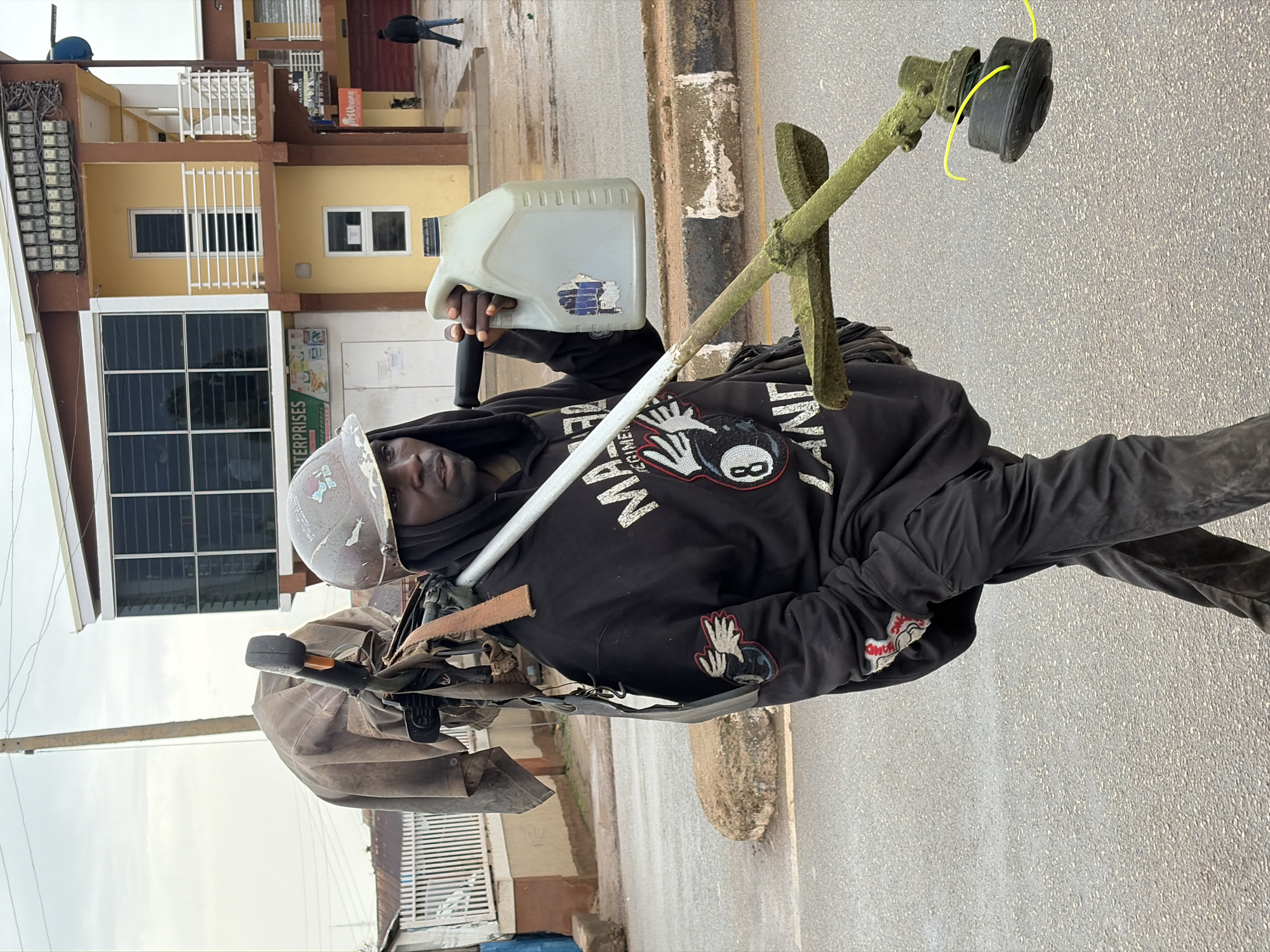 A Black man in a hard hat and black hoodie carries a string trimmer over his left shoulder, a plastic jug in his right hand. He walks on a paved street in Jos, Nigeria, with a building and power lines in the background. The man has dark skin, black hair, and is wearing gloves. The string trimmer appears to be gas-powered, with a green shaft and black trimmer head. The jug is white, opaque, and has a black cap. The building in the background is yellow and brown, with several windows and balconies. Power lines are visible overhead. The street is wet, suggesting recent rain.