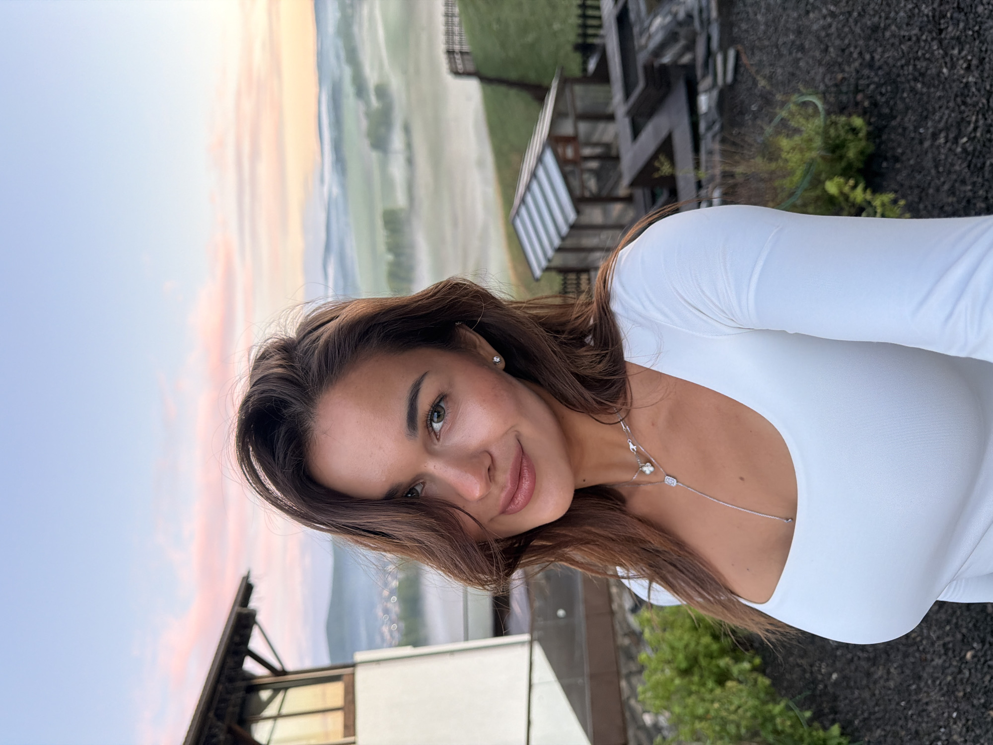 A young woman is posing for a selfie outdoors, wearing a white, long-sleeved top and a delicate silver necklace. Her dark brown hair is loosely styled, and she is smiling slightly at the camera. Behind her, a landscape of rolling hills is visible, partly obscured by mist or fog. The sky is illuminated with soft hues of pink and blue, indicating sunrise or sunset. Architectural details, including wooden structures and a dark, gravelly ground, suggest a residential or resort setting. Based on the prompt, the location is potentially in Shamoni, Russia.