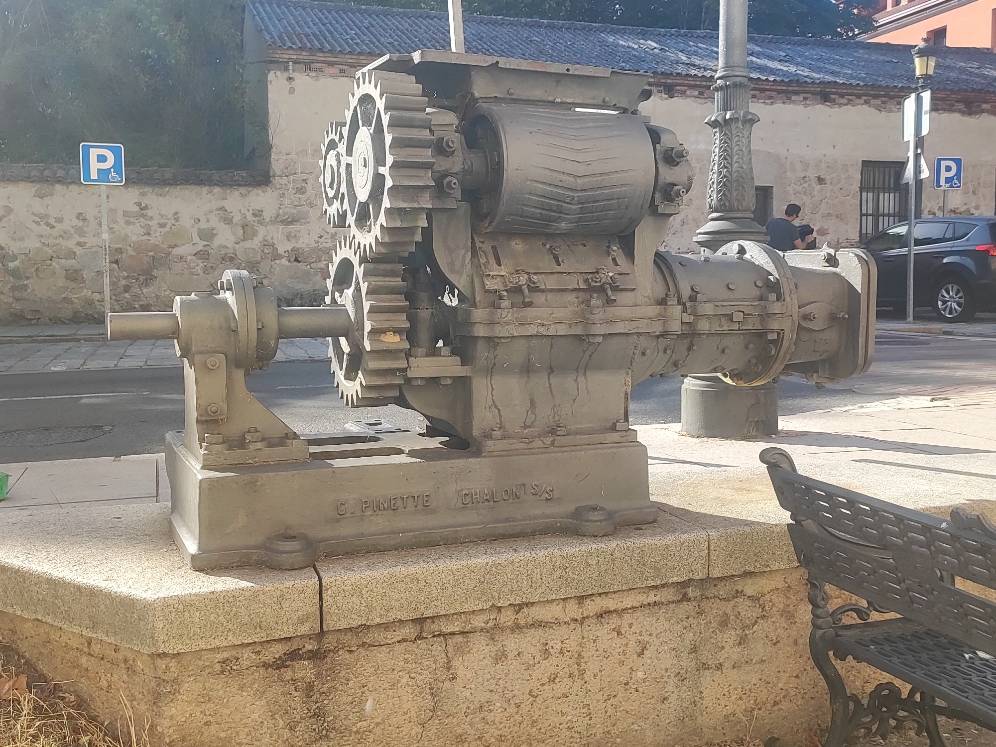 The image shows a large, old industrial machine with gears, situated on a stone pedestal in an outdoor urban environment. The machine is dark gray and appears to be made of metal. Text is visible on the base of the machine, reading "G. PINETTE CHALONS/S". Behind the machine, a stone building with a tiled roof is visible, along with a street and a parking sign. A black car is parked on the street. A black wrought-iron bench is positioned to the right of the machine. The scene appears to be in the city of San Ildefonso, Spain.