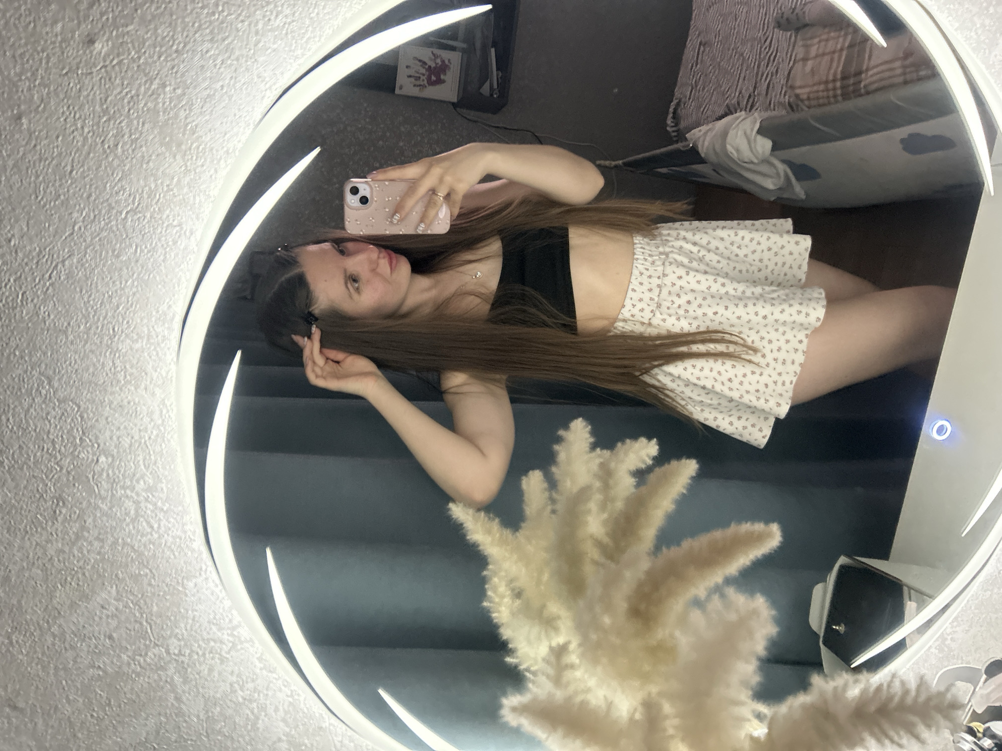 A young woman is taking a selfie in a mirror. She is wearing a black crop top and a white floral skirt. Her hair is long and brown. The background is a grey wall and a bed. There is also a decorative object in the foreground. The location is not evident in the image.